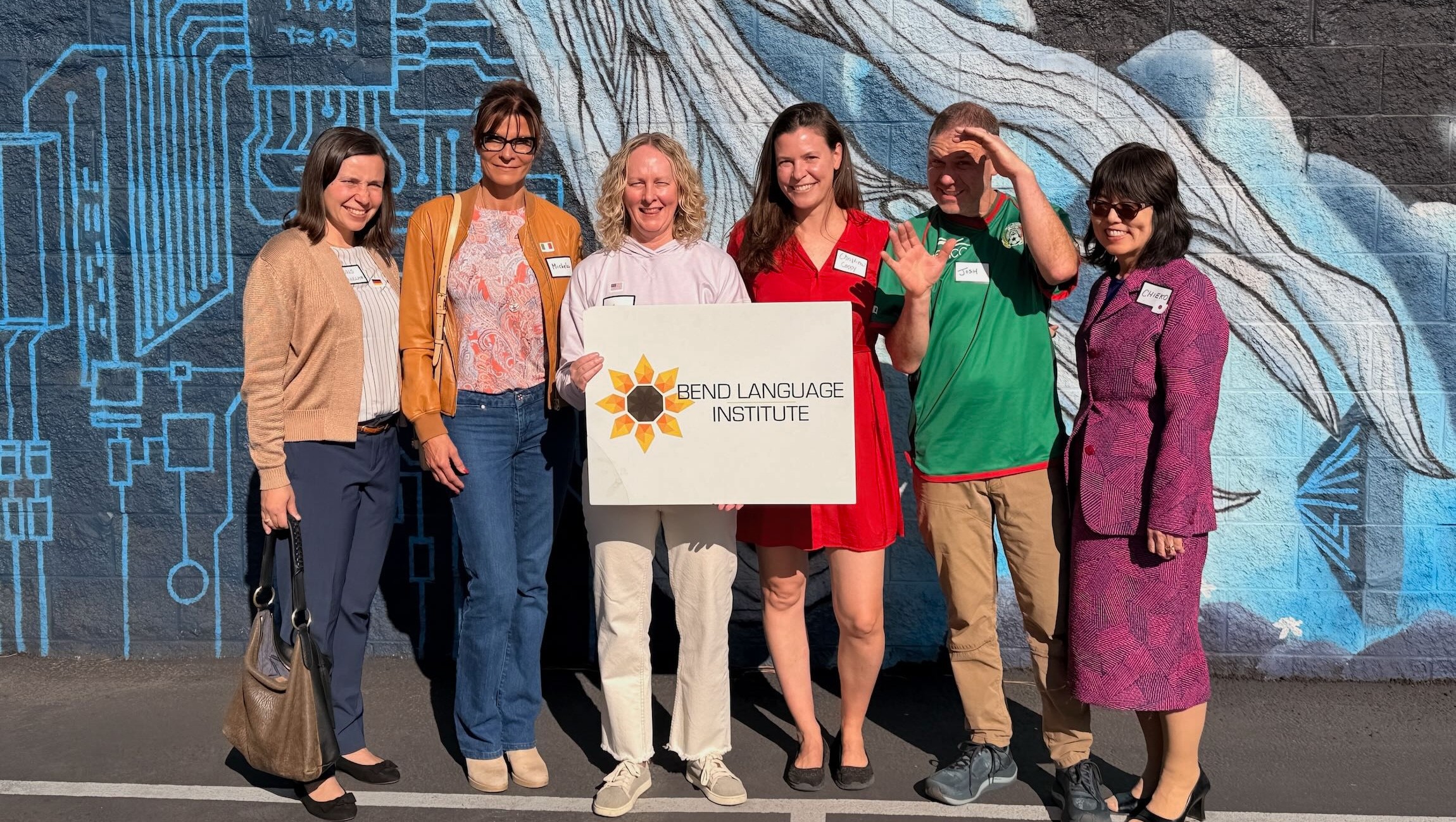 Group of seven women standing outdoors in front of a mural, holding a sign with a sunflower logo and text.