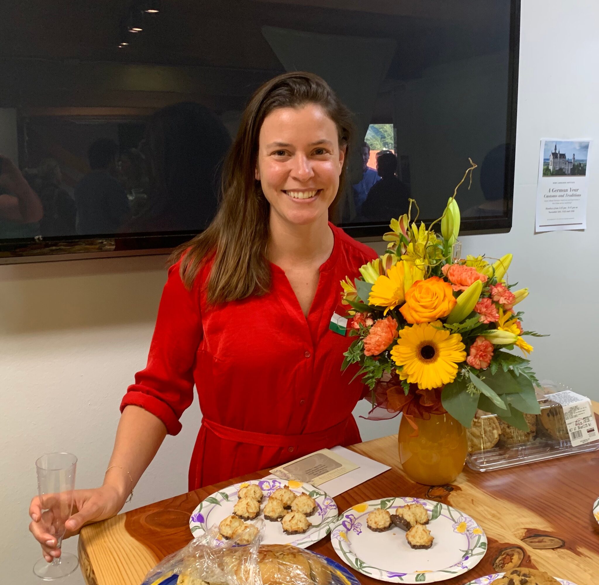 Woman in red dress holding a bouquet of yellow and orange flowers, smiling, standing behind a table with cookies and plates.