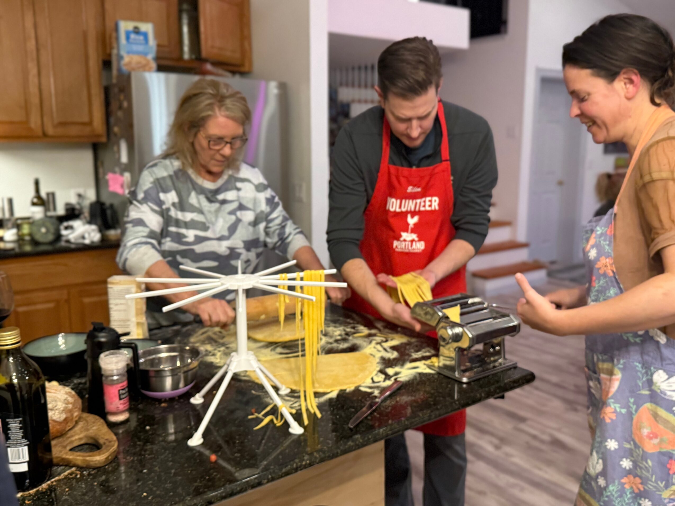 Three people in a kitchen making pasta, with a pasta machine and dry pasta on the counter.