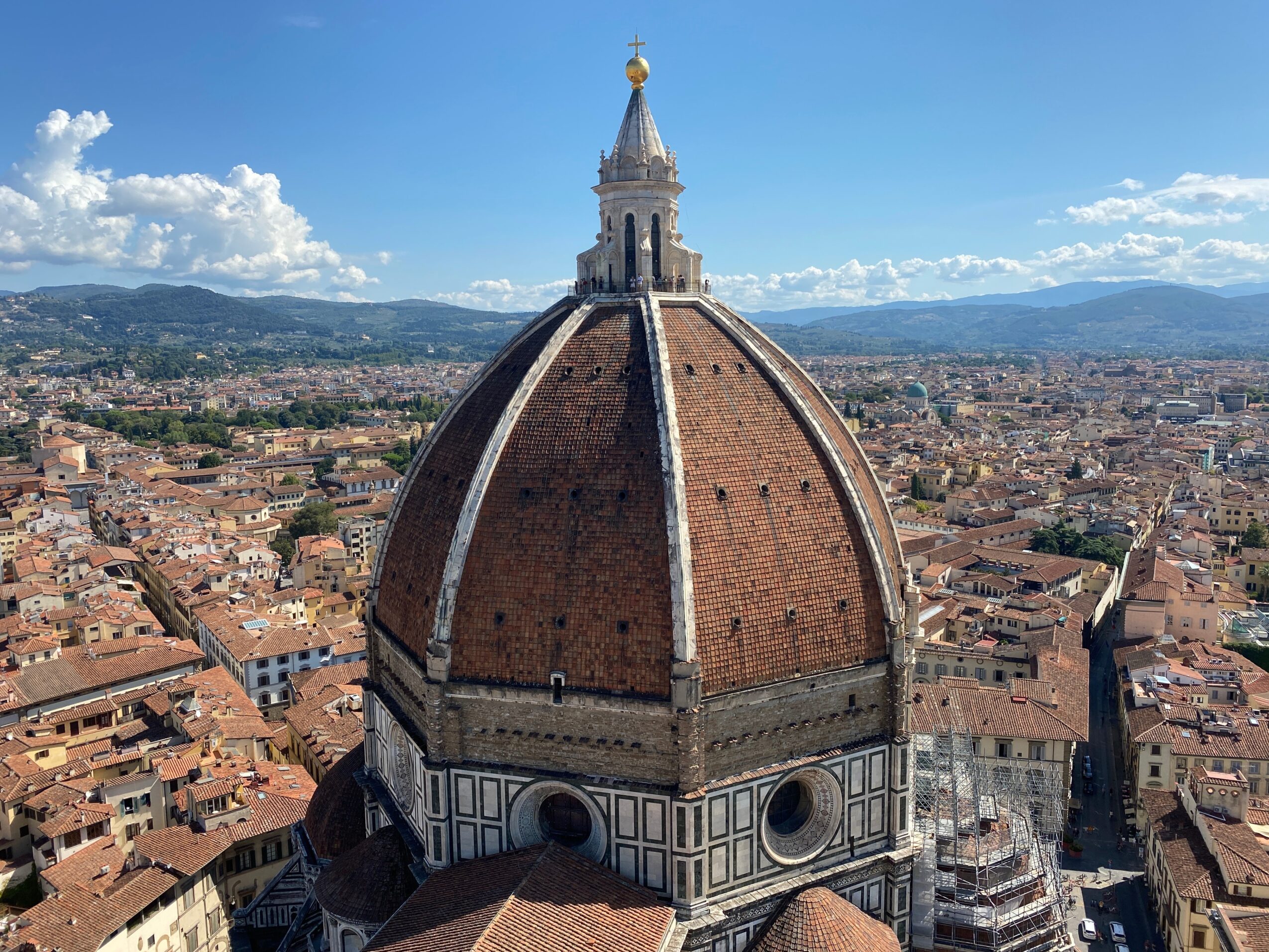 Dome of Florence Cathedral with cityscape and mountains in background, blue sky with clouds.