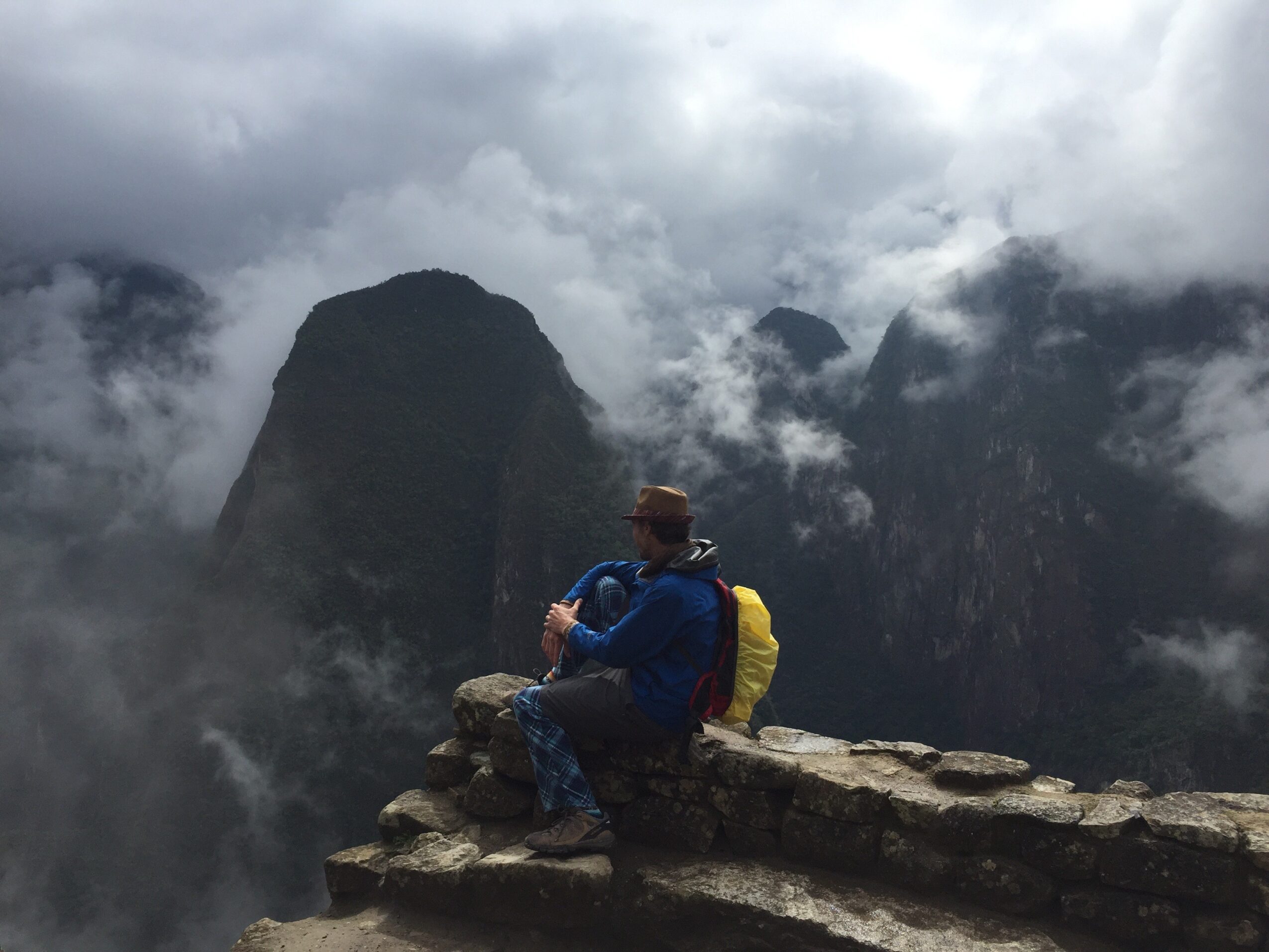 Person sitting on rocks with mountains and clouds in the background, wearing a hat and backpack.