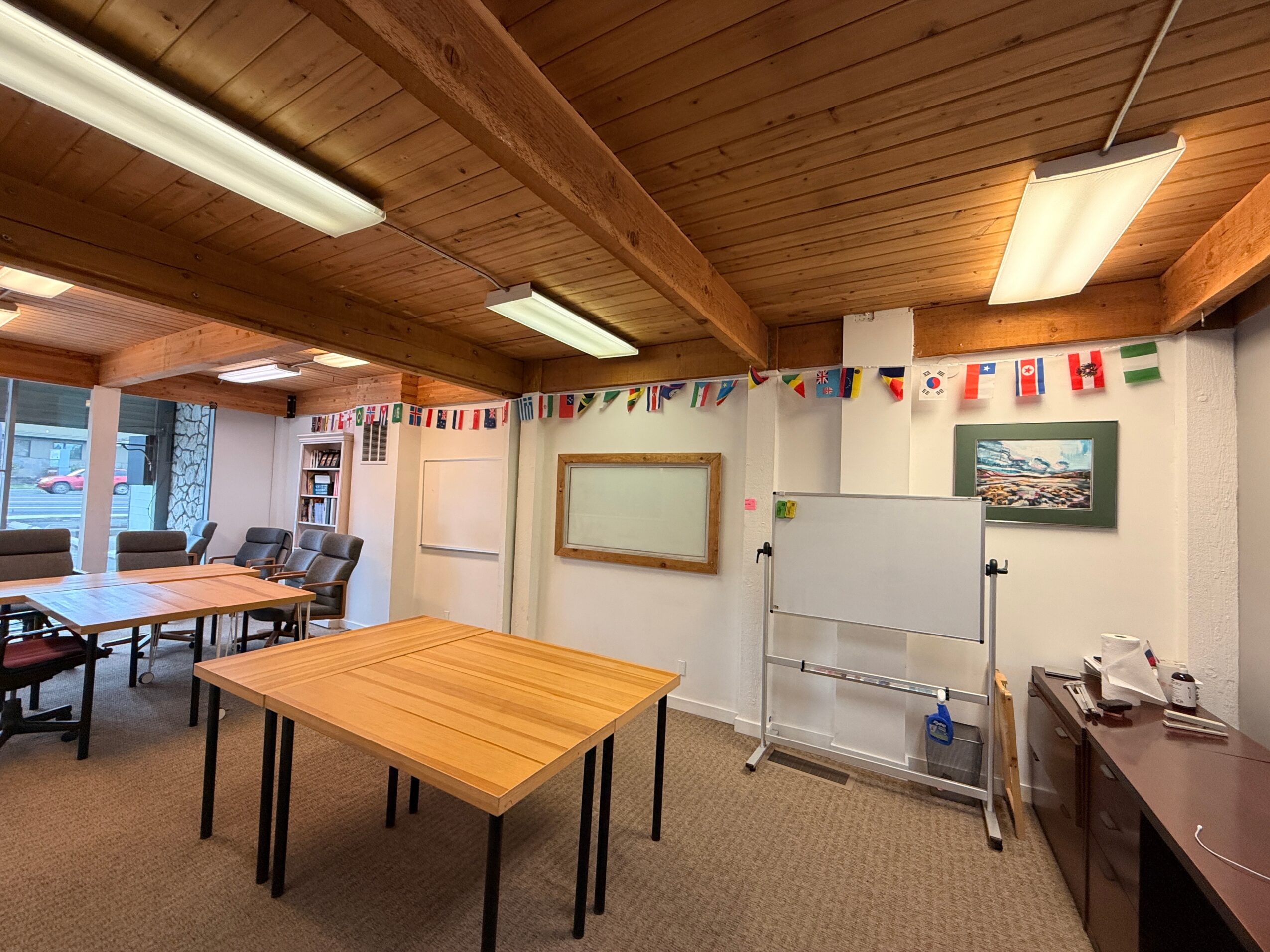 Classroom with wooden ceiling, tables, whiteboard, and decorations, including a banner of international flags, and a window with a view outside.