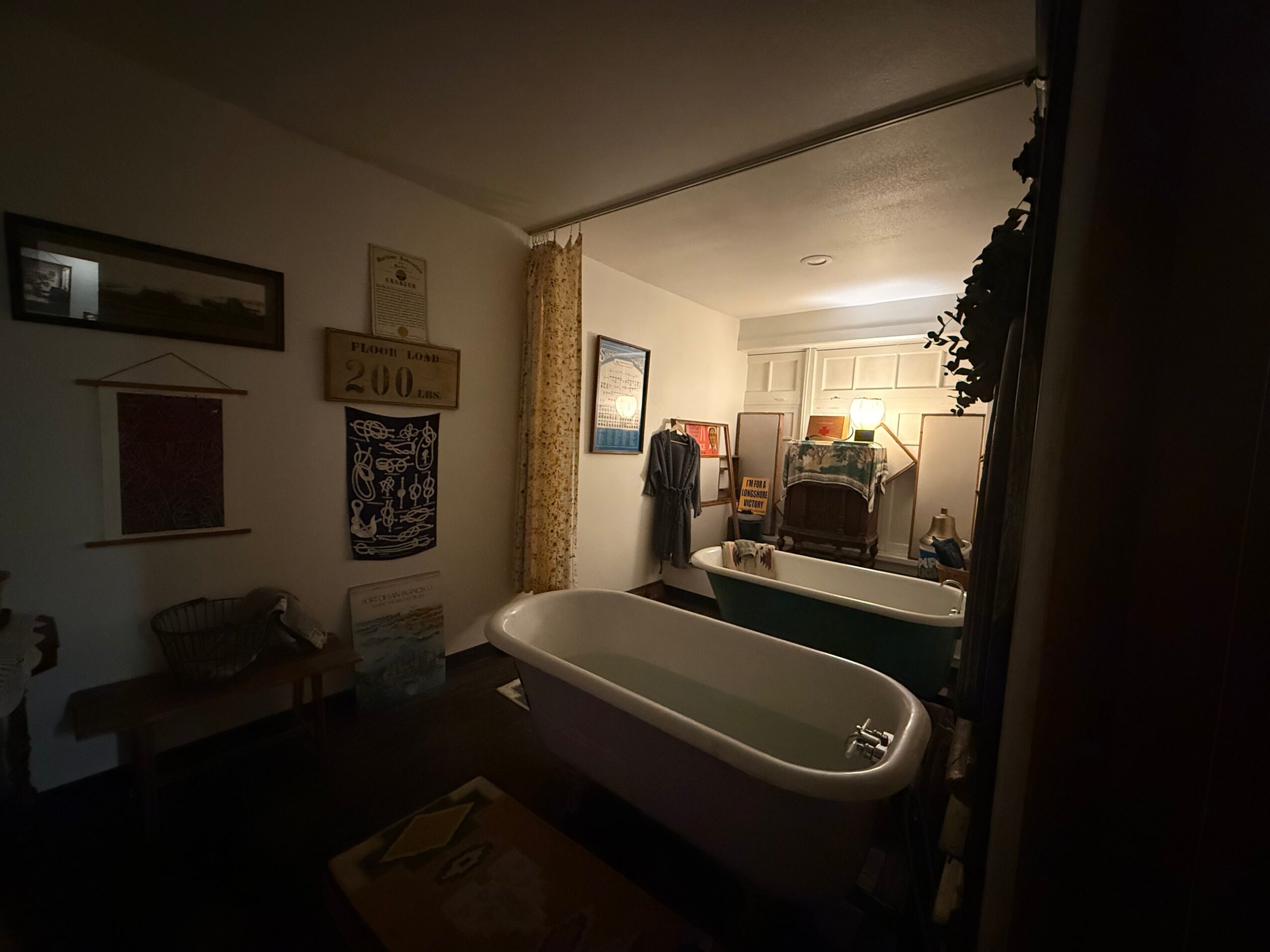 Bathroom with a bathtub, towels, artwork, and a window with curtains, illuminated by a lamp, viewed from the doorway.