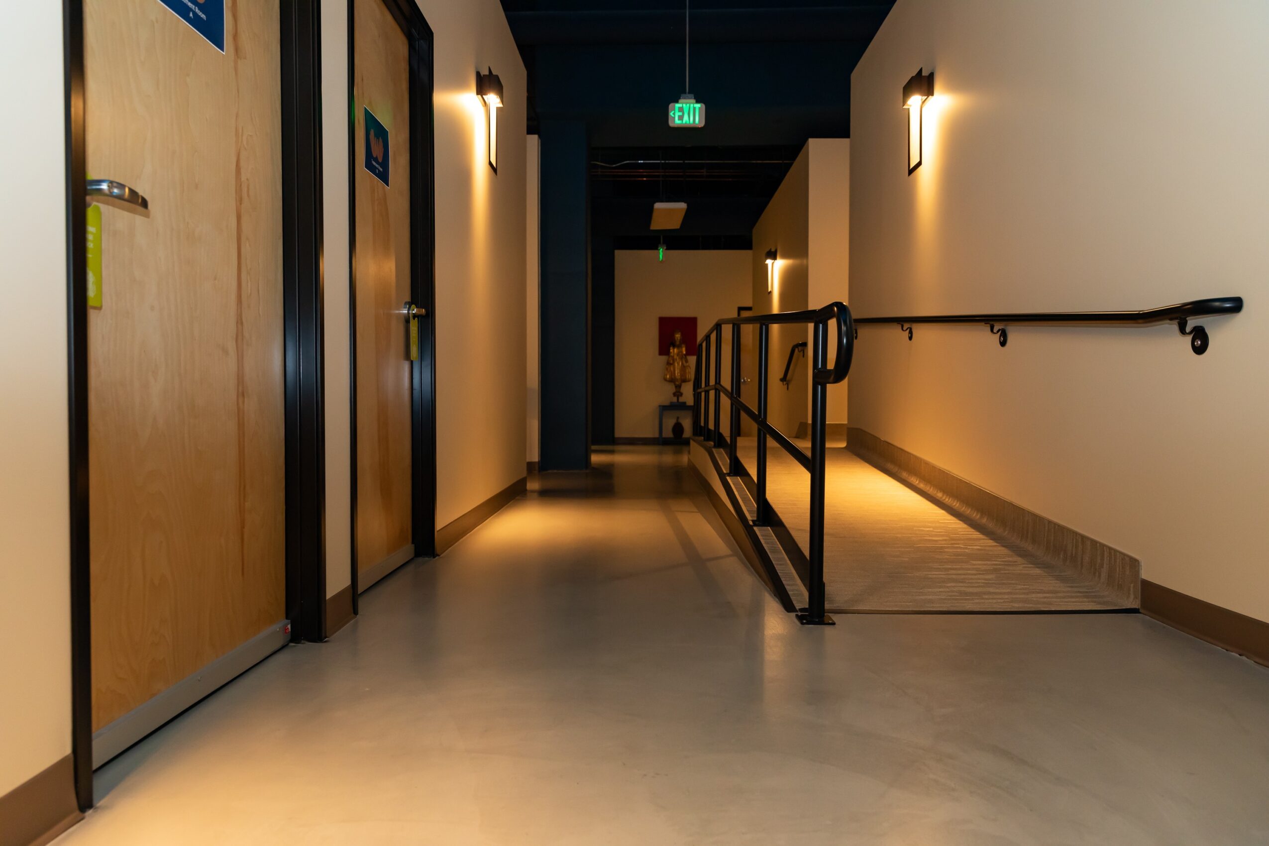 Hallway with elevator doors on the left, handrails on the right, and a door at the end, lit by wall lights.