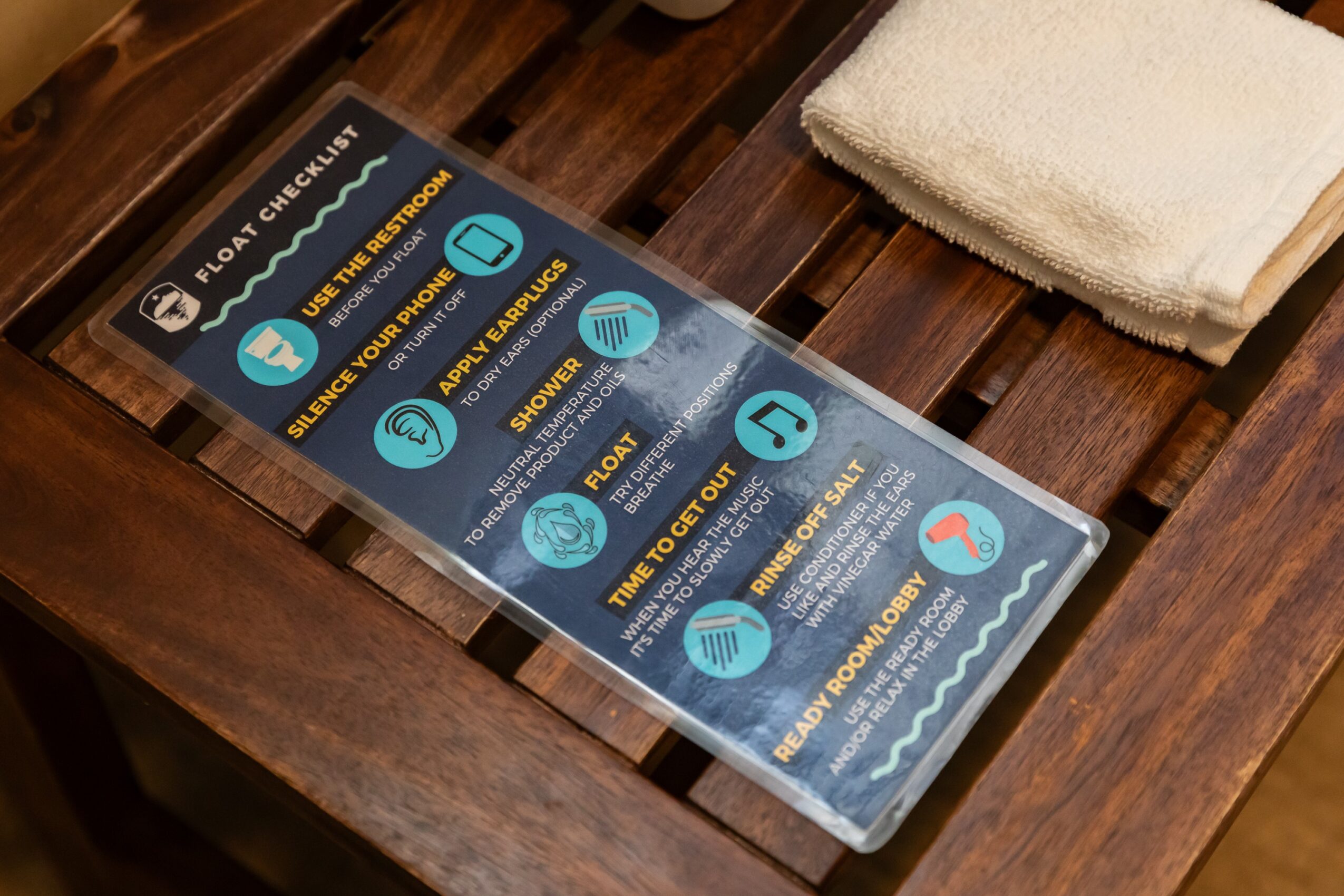 A clear acrylic sign with blue icons and text about a facility's features, placed on a wooden surface with a folded towel nearby.