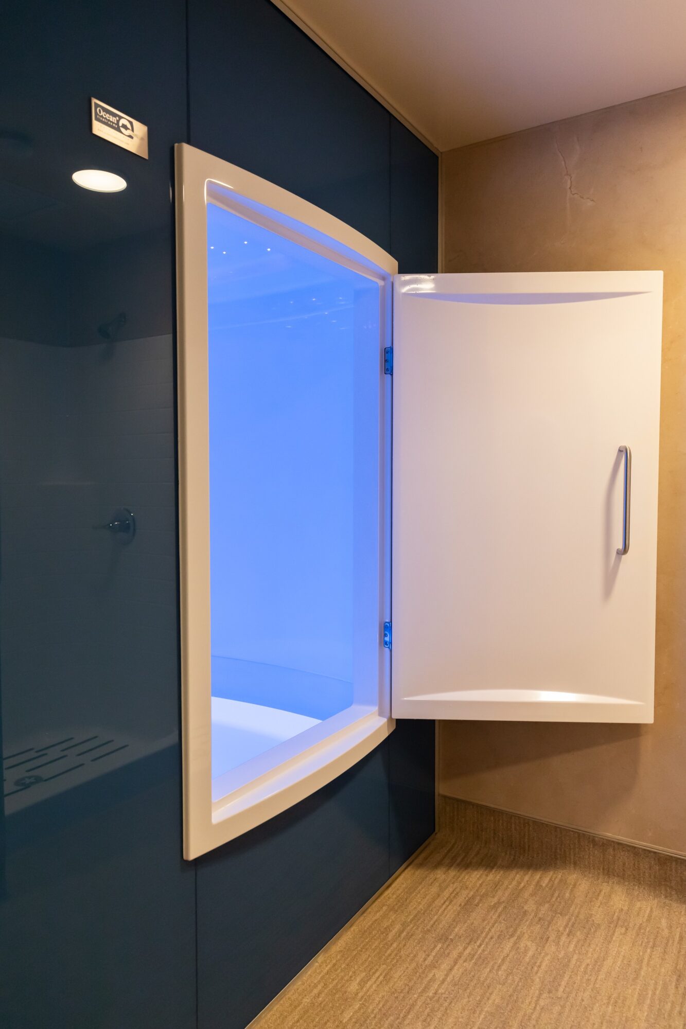 A white wall-mounted cabinet with a door open, revealing a blue-lit interior, against a dark wall with a beige wall nearby.