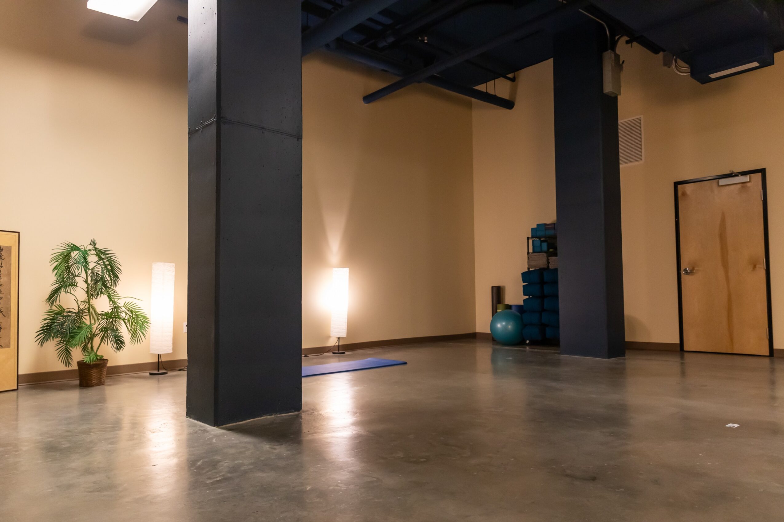 Empty room with black columns, a potted plant, a blue vase, and a door, with beige walls and a polished floor.