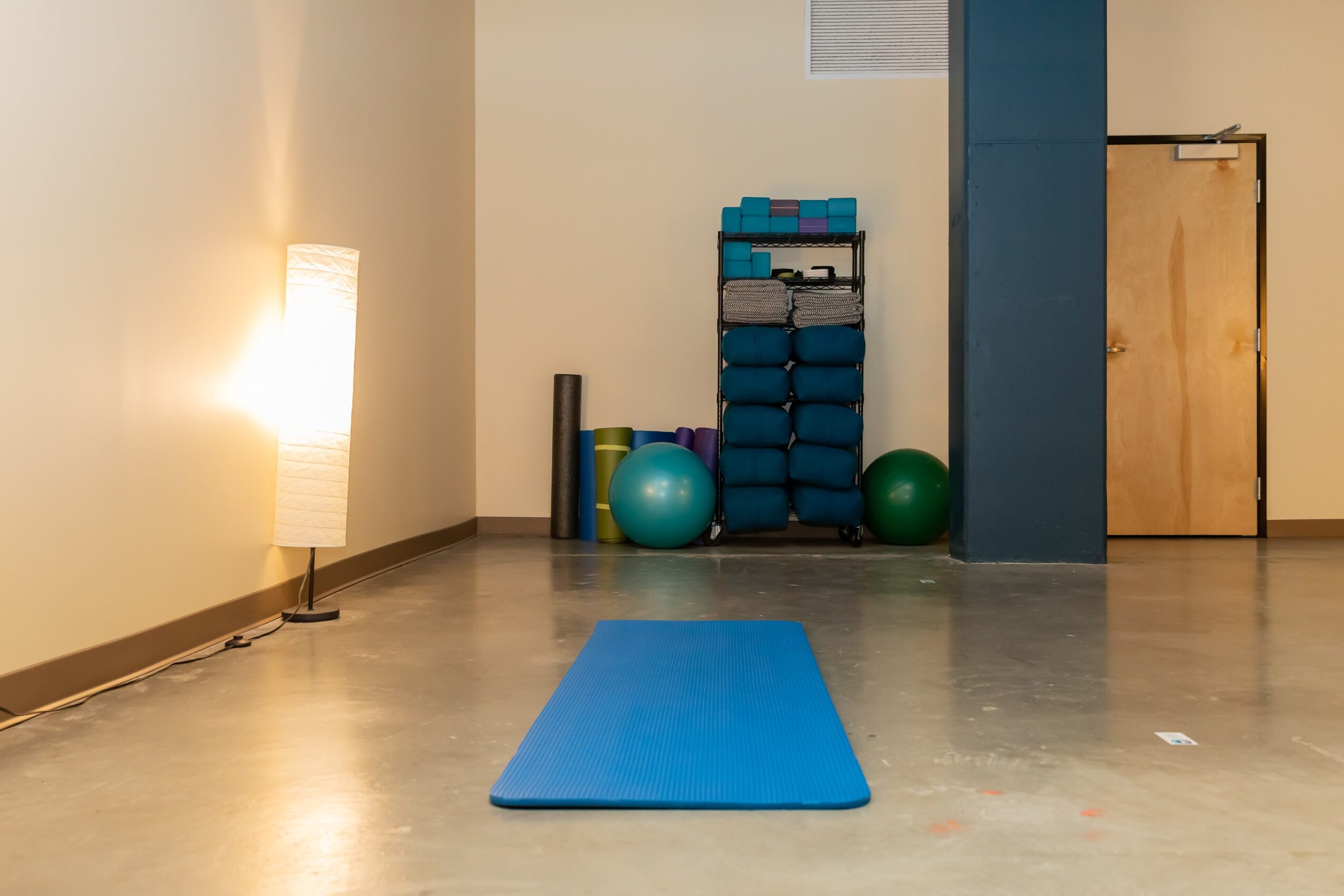 Empty room with a blue yoga mat, exercise balls, and a storage rack against the wall, lit by a floor lamp.