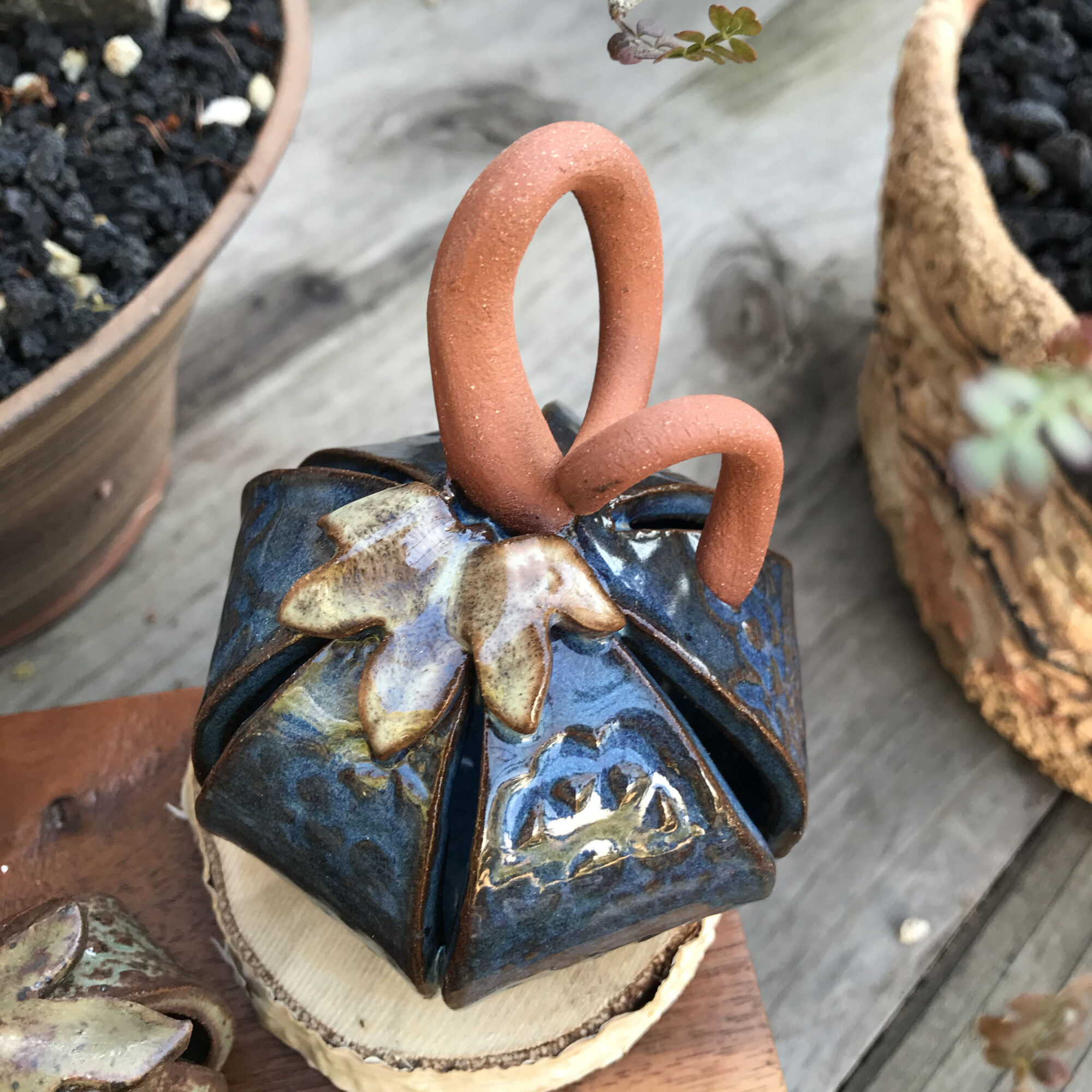 Decorative ceramic object resembling a gift box with a handle, placed on a wooden surface with potted plants nearby.