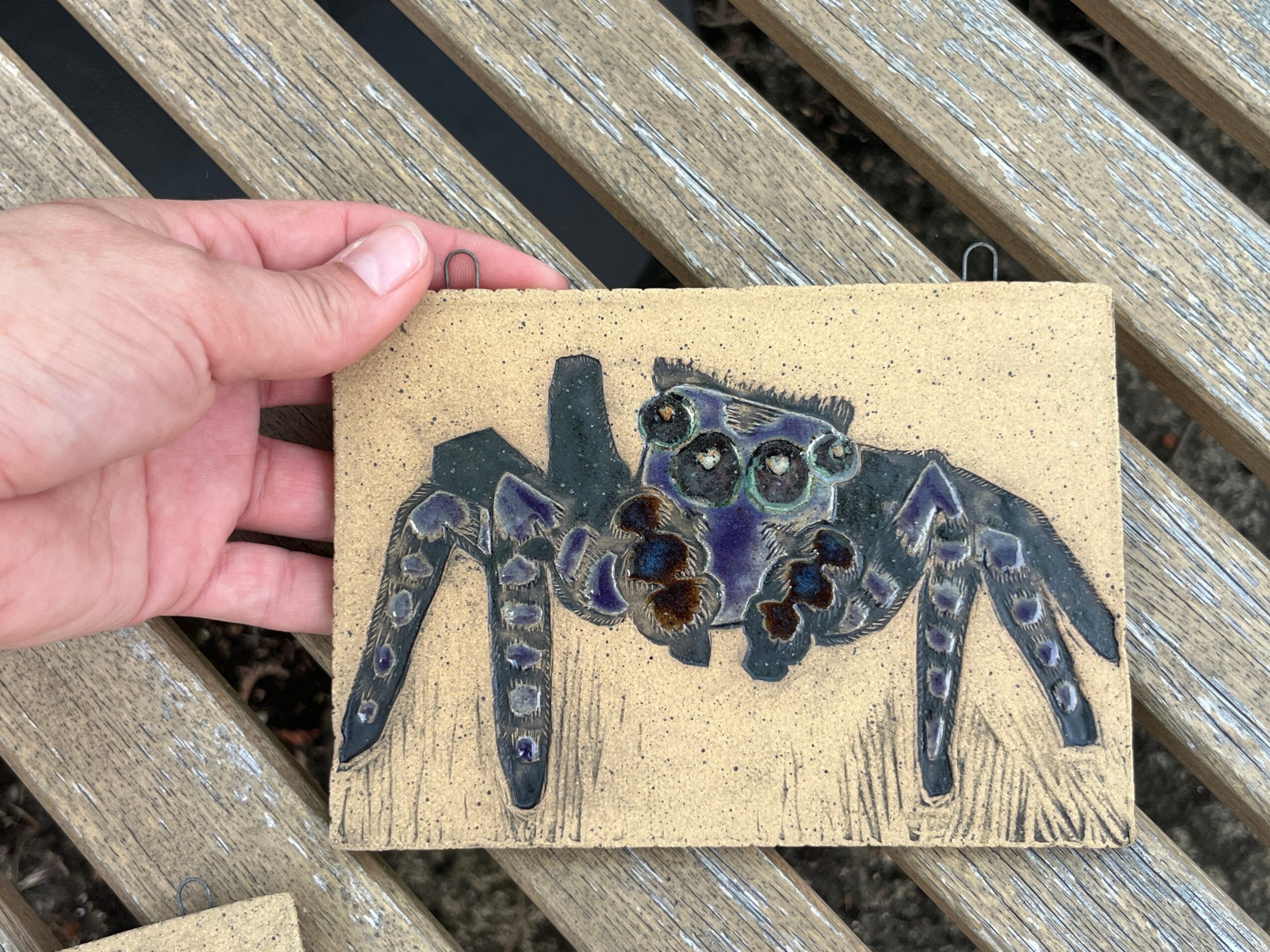 Hand holding a small rectangular artwork of a blue and black spider on a beige background, placed on a wooden surface.
