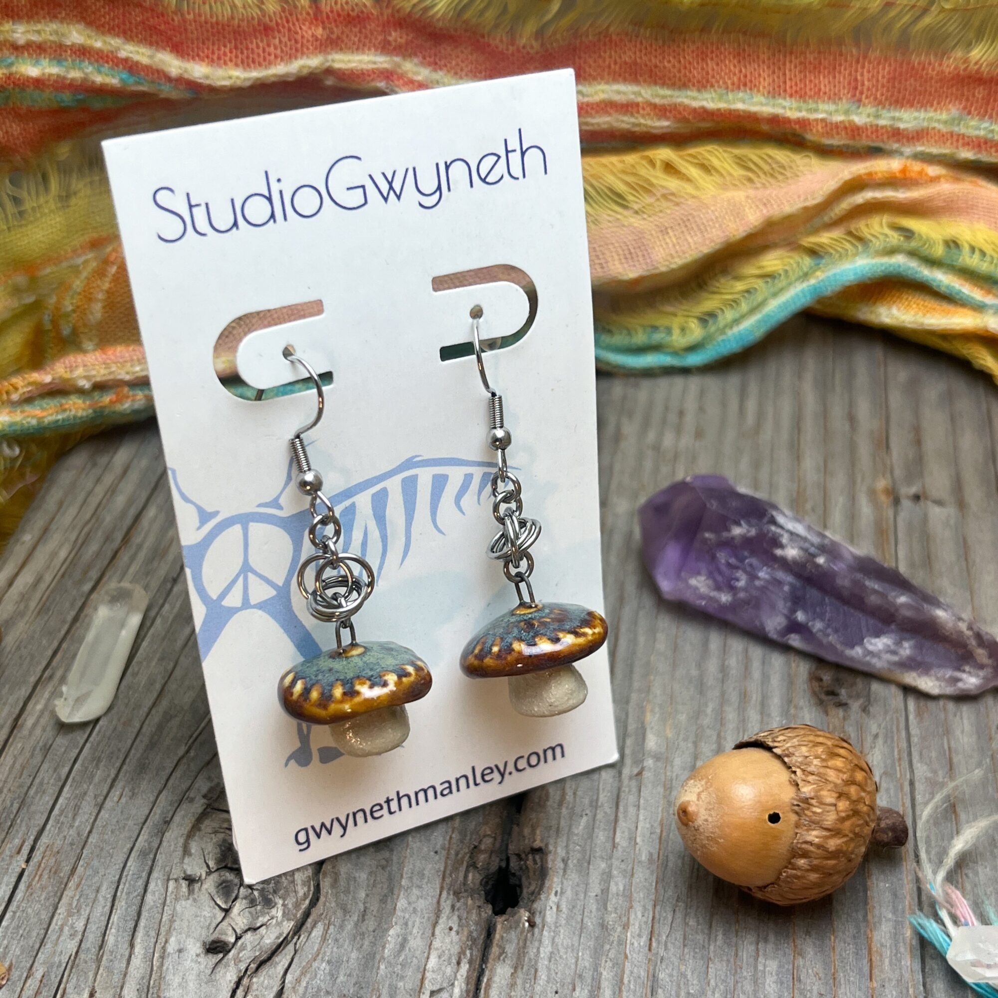 Pair of earrings with mushroom charms on a display card, placed on a wooden surface with a purple crystal and a small wooden figure nearby.