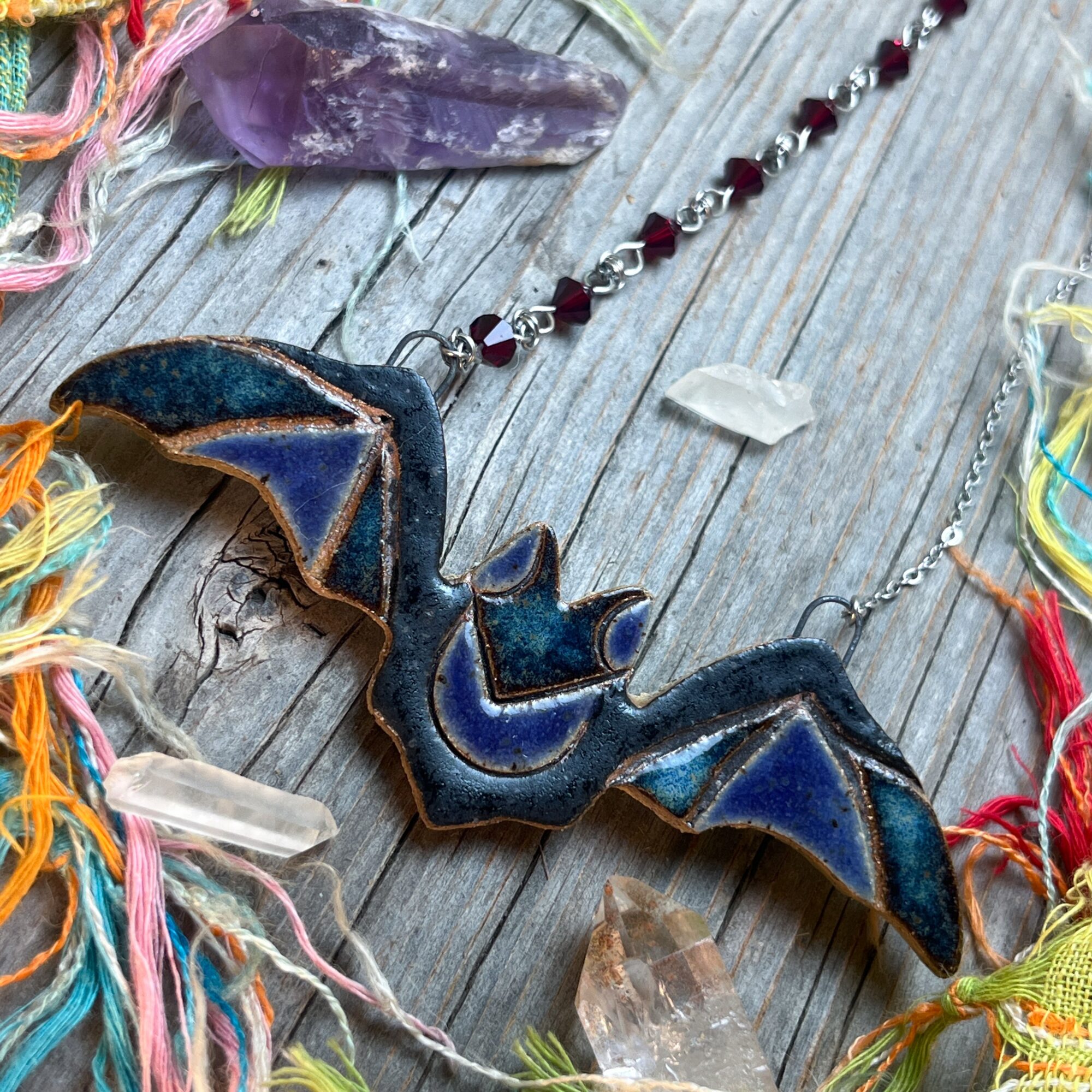 Blue bat-shaped pendant with a chain, surrounded by colorful yarn and crystals on a wooden surface.