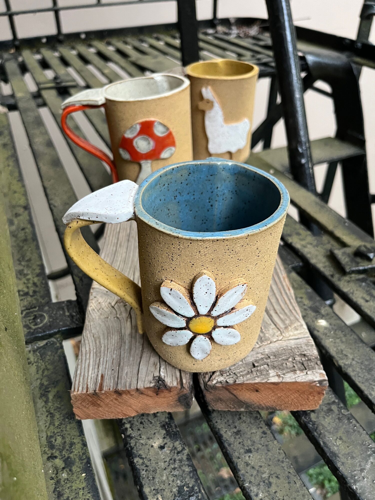 Three decorated cups with flowers and mushrooms are placed on a wooden bench outdoors.