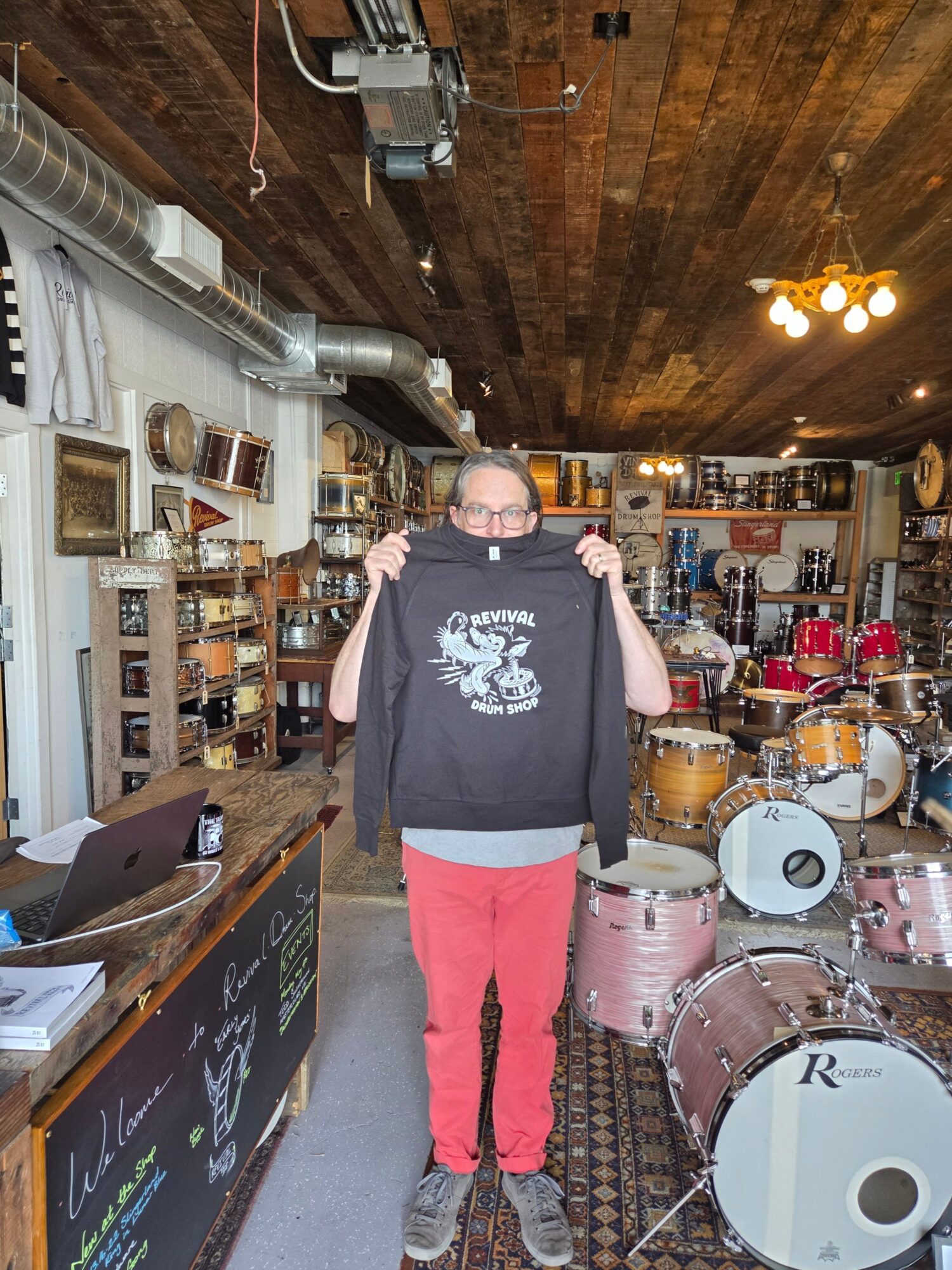 Person standing in a music store with drums and musical instruments, holding a guitar, wearing glasses, black t-shirt, and red pants.