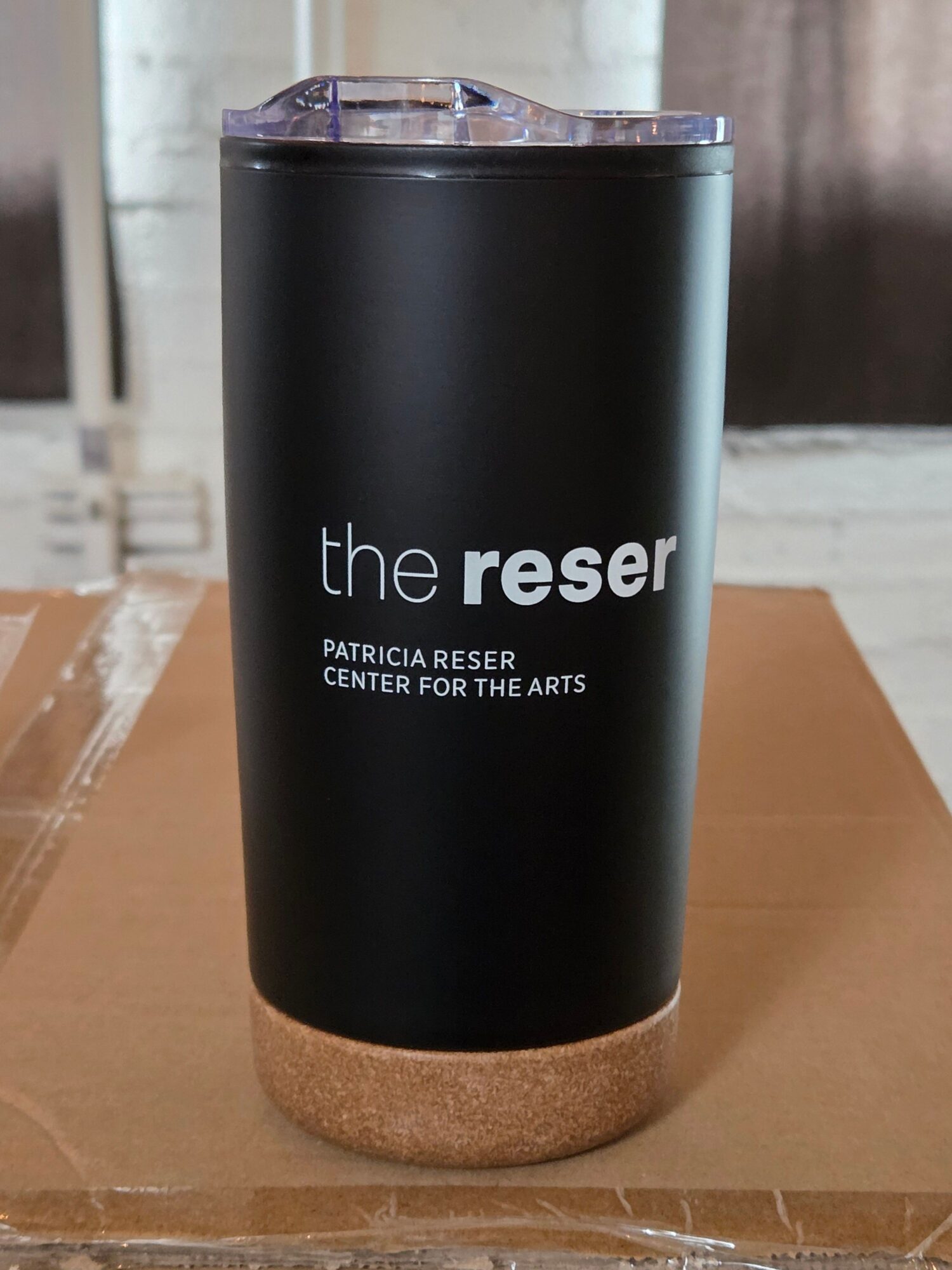 Black tumbler with white text reading 'the reser' and additional smaller text, placed on a cardboard surface.