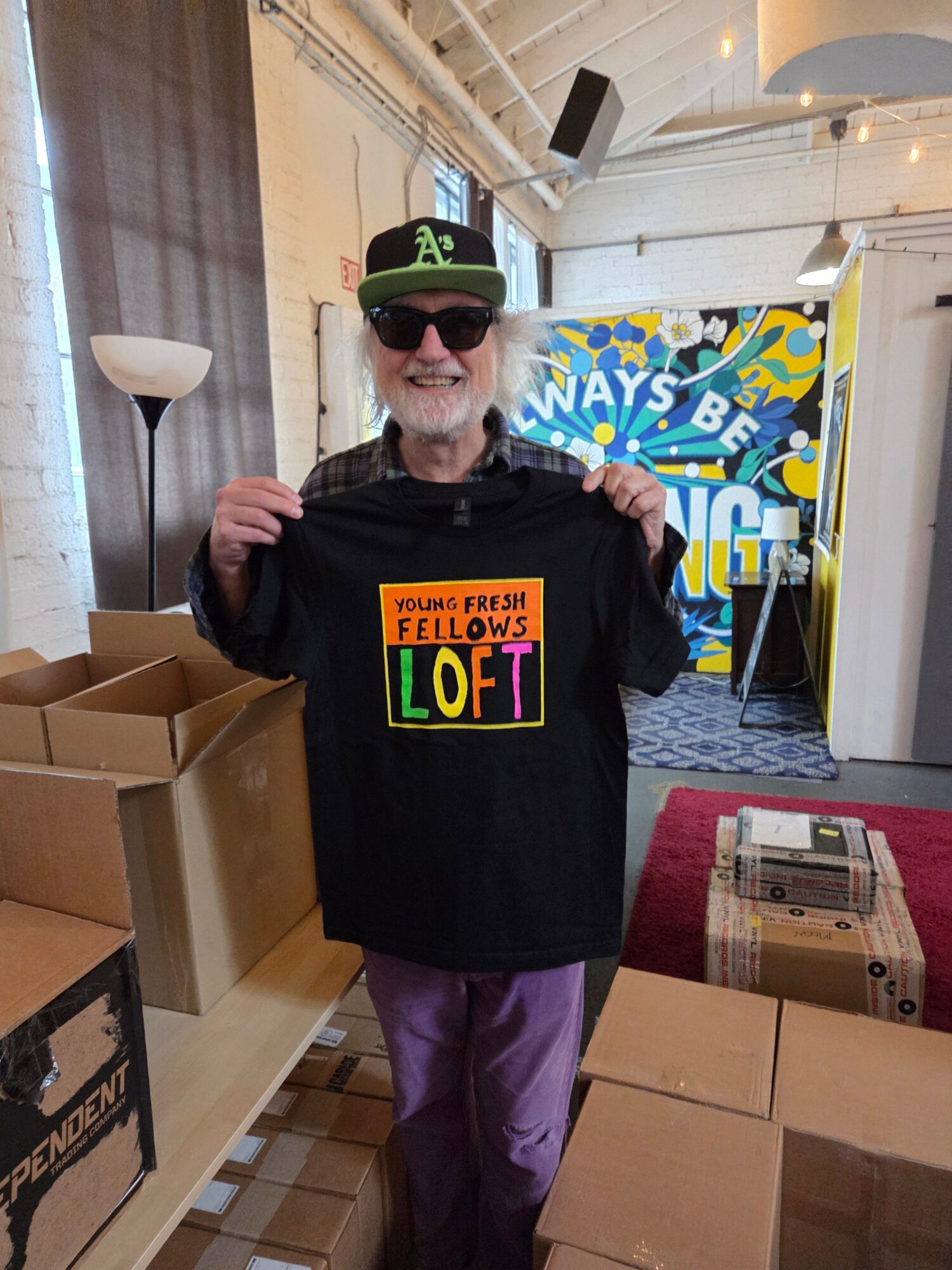 Person wearing a black hoodie with colorful text, black cap, and sunglasses, standing indoors among boxes and a lamp.