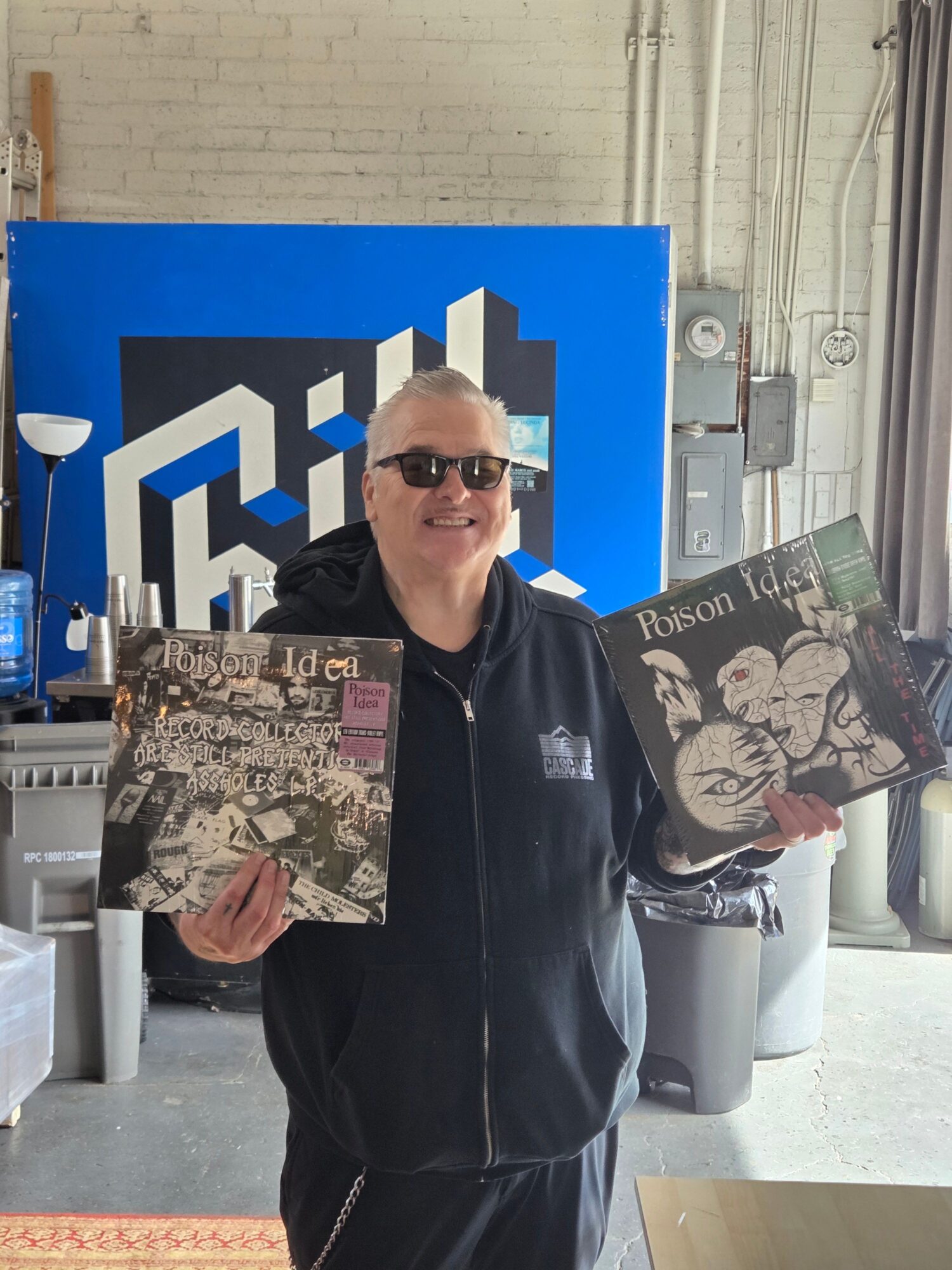 Person holding two vinyl records in front of a blue and black sign, wearing sunglasses and a black jacket.