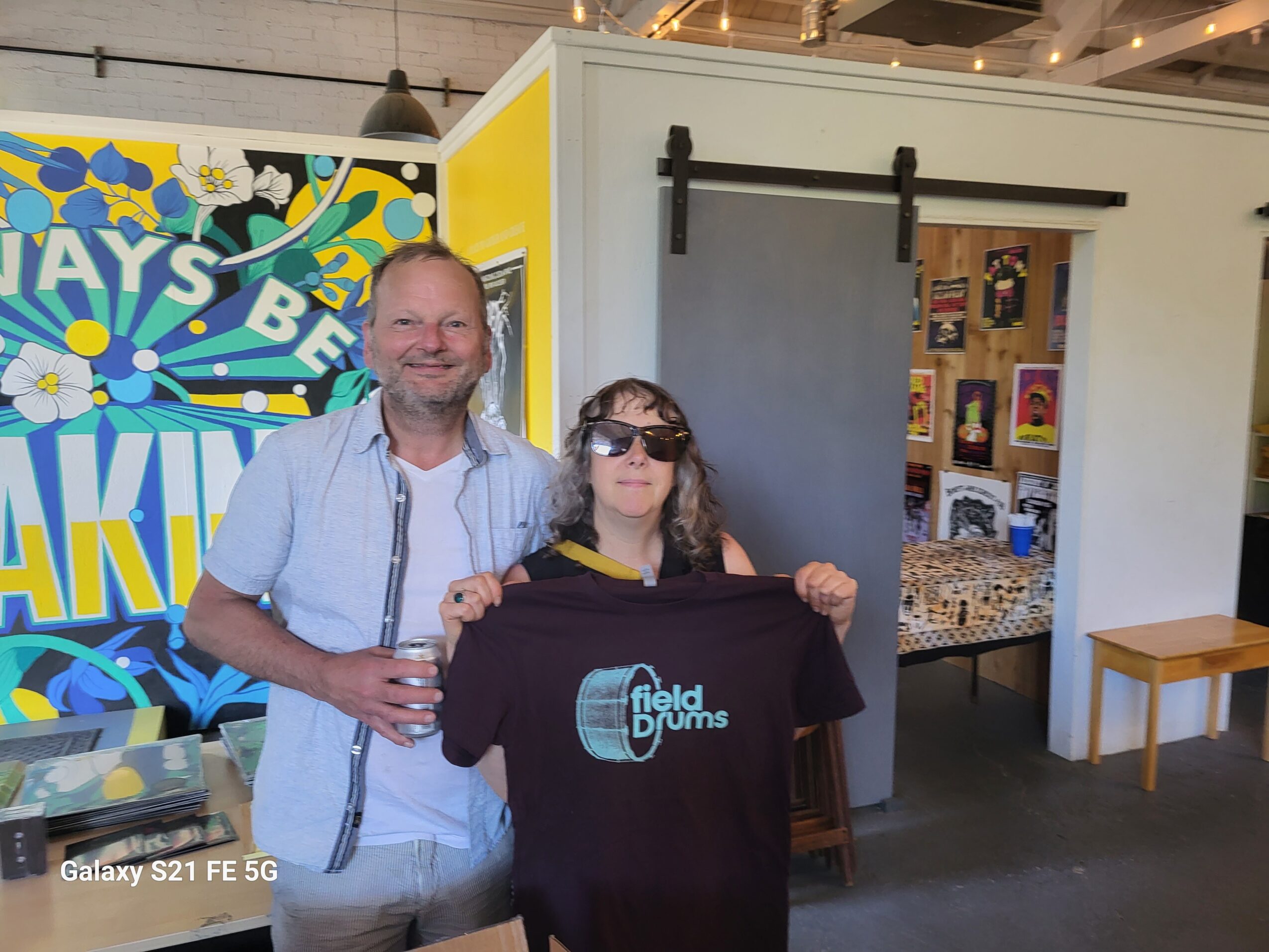 Two people standing indoors, one holding a black T-shirt with a logo, smiling, with a colorful background and a sliding door behind them.