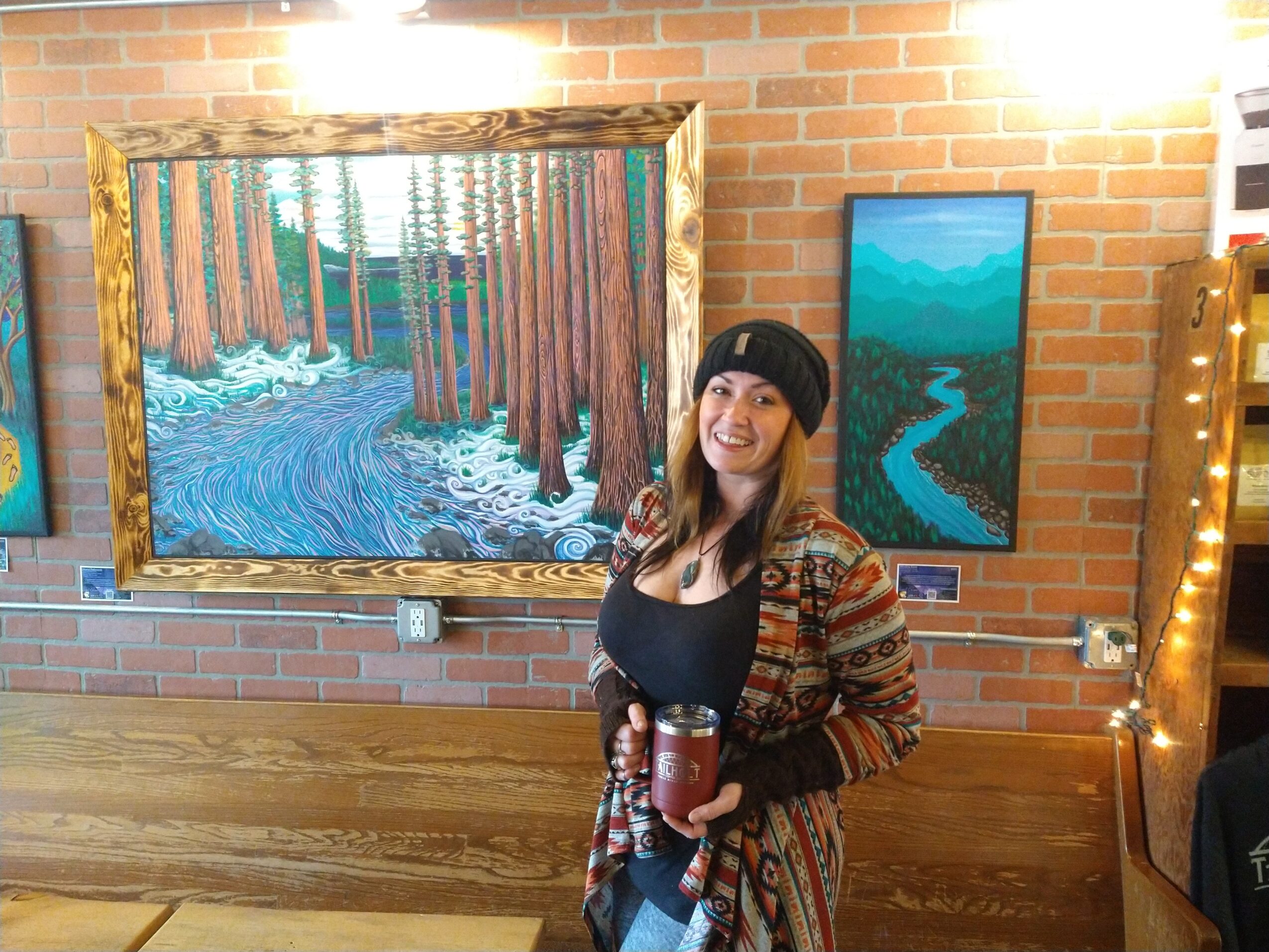 Woman standing in front of two landscape paintings, holding a drink, smiling, brick wall background.