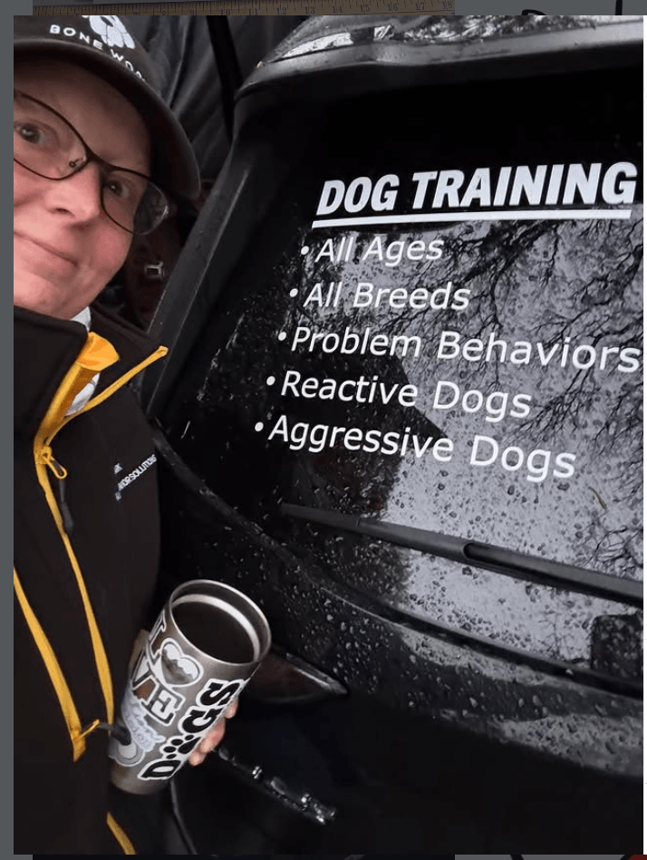 Person wearing glasses and a hat holding a travel mug, standing next to a sign about dog training for all ages and breeds.