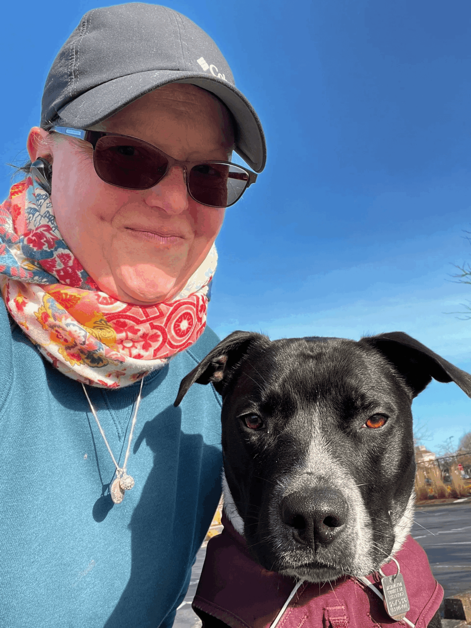 Person wearing sunglasses, a gray cap, and a colorful scarf, smiling with a black and white dog outdoors against a blue sky.