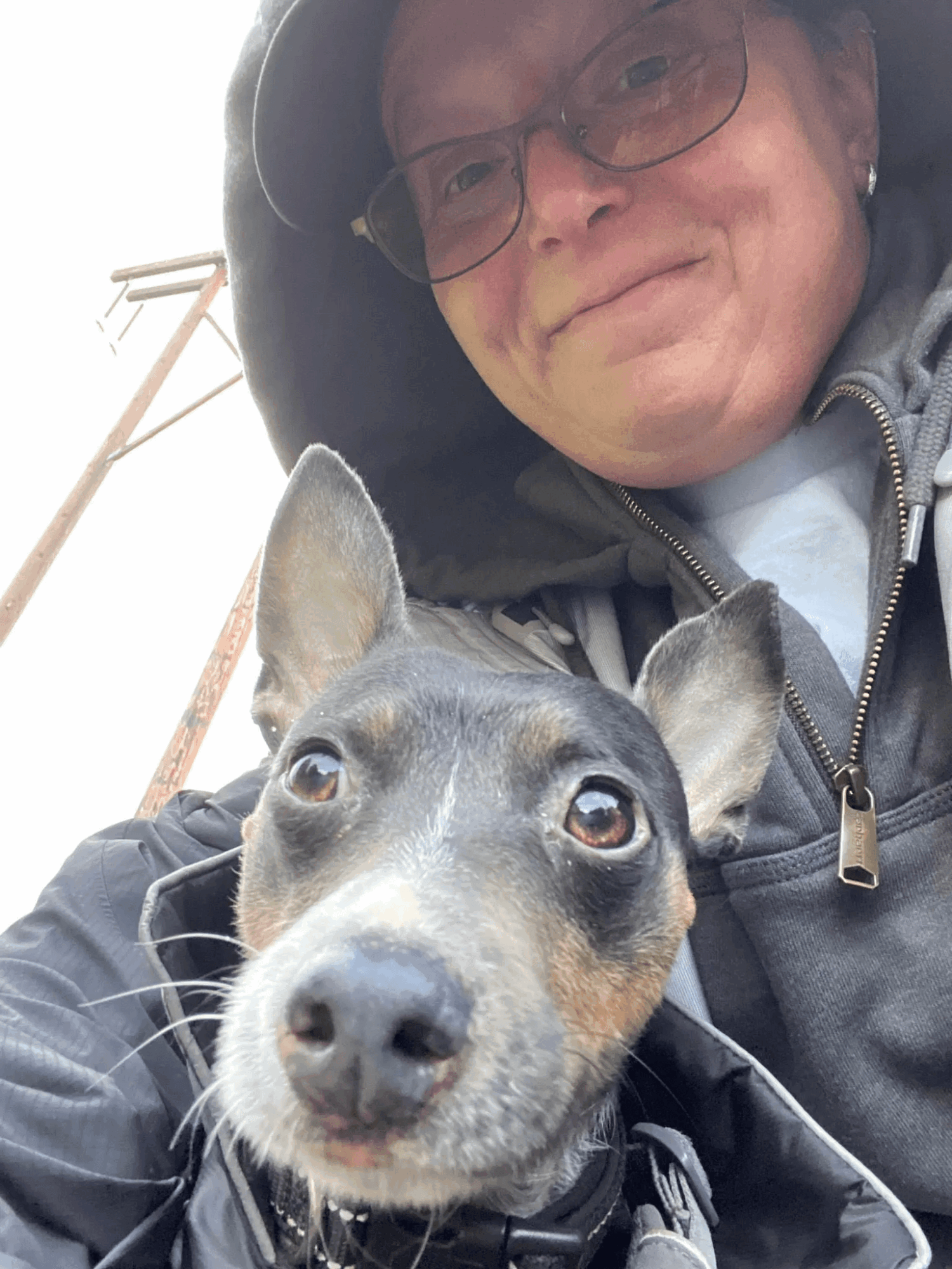 Person wearing glasses and a hooded jacket taking a selfie with a dog outdoors.