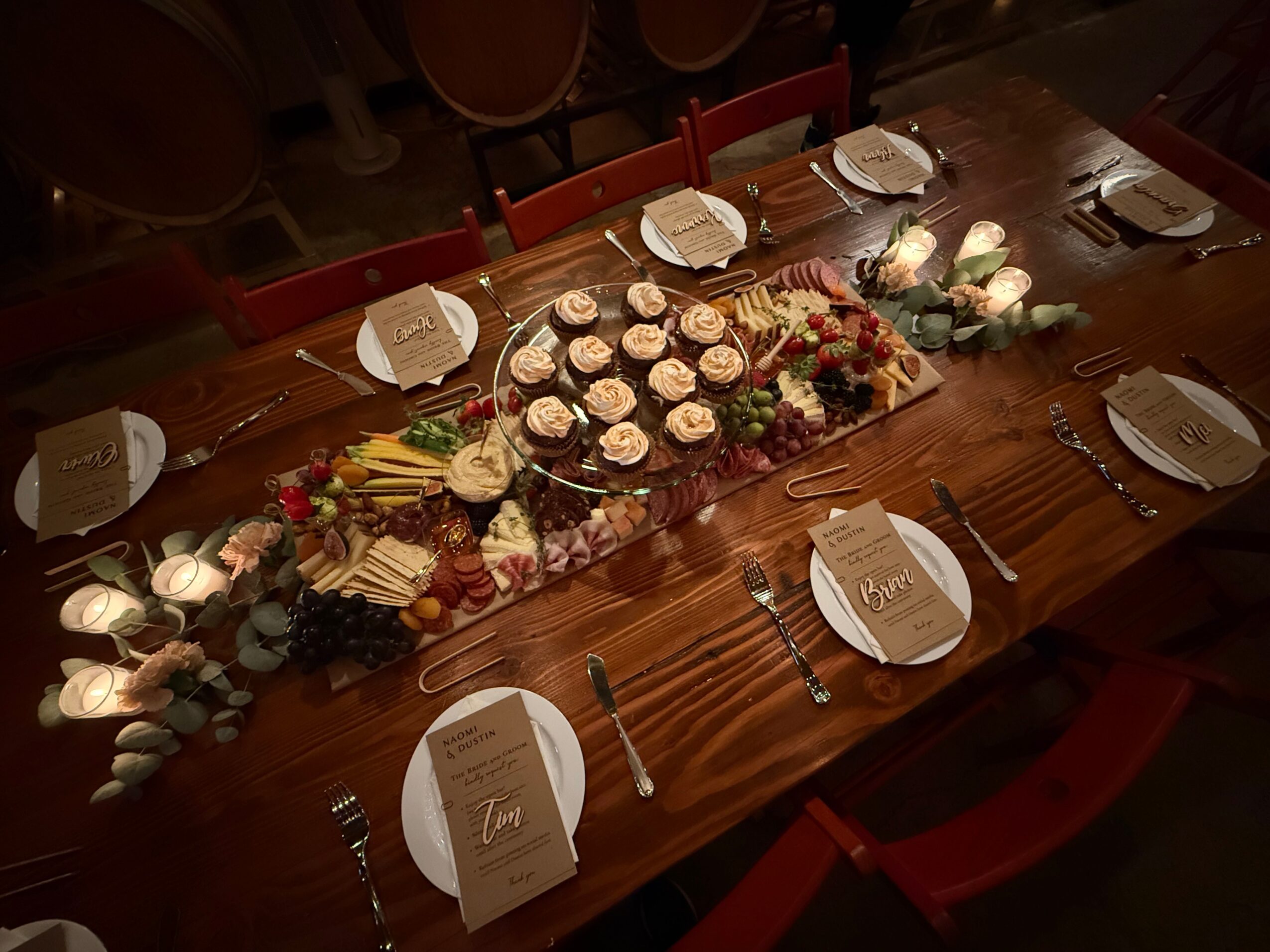 Long wooden table set for a celebration with plates, utensils, and a central dessert platter with cupcakes and fruit, illuminated by candles.