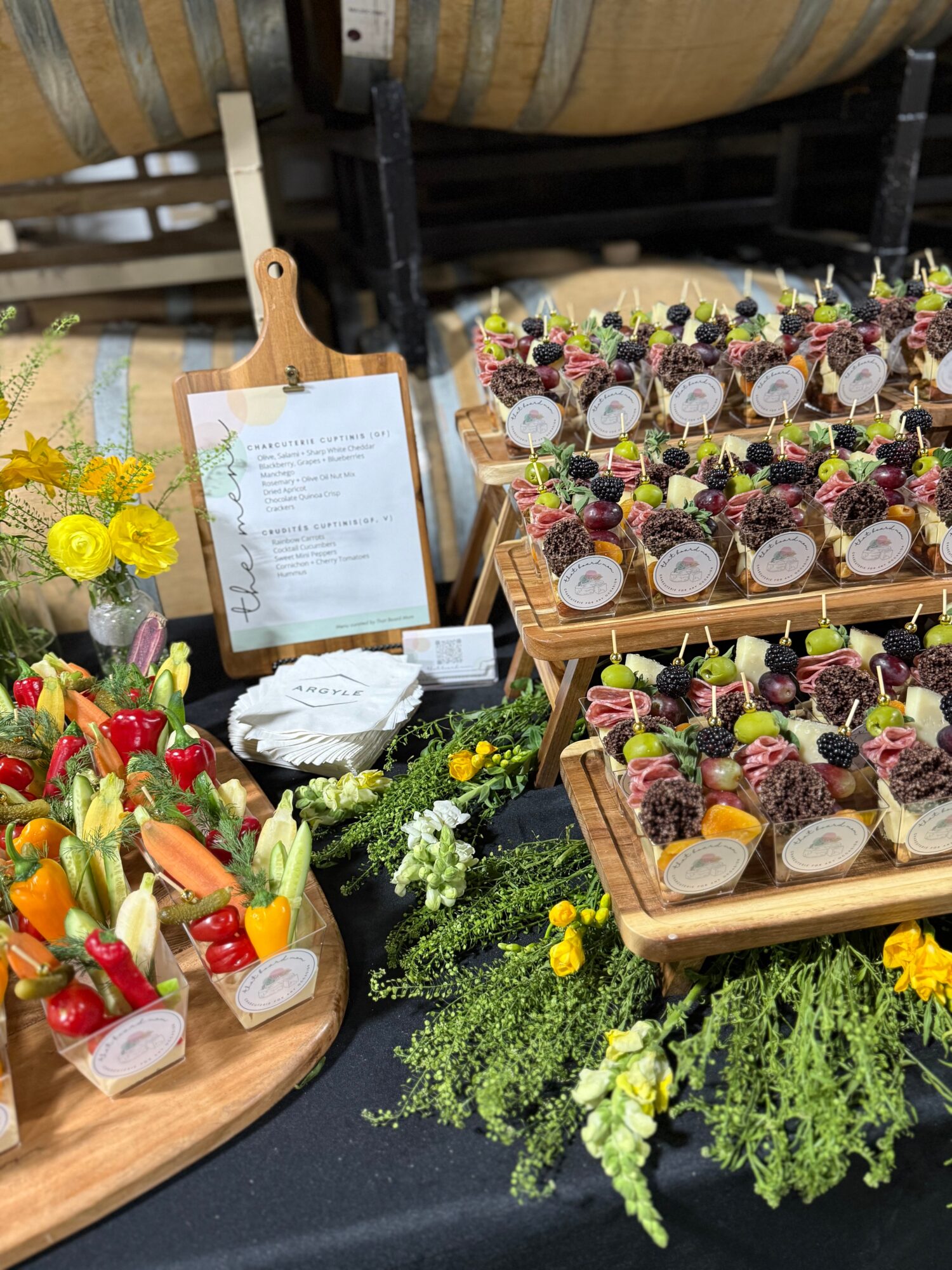 Display of colorful fruit and flower arrangements with a menu on a wooden stand, set on a black table with greenery.