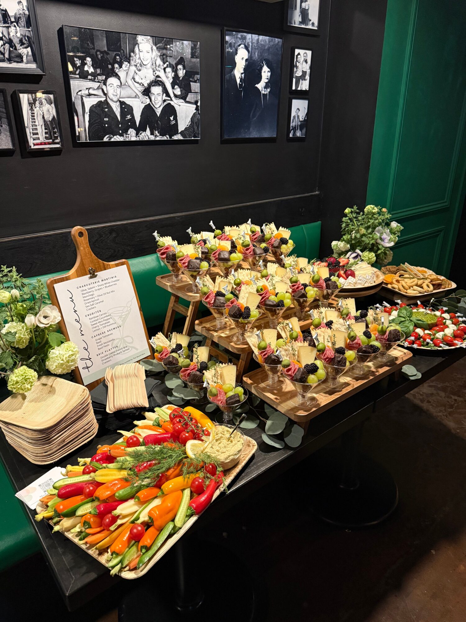 Table with colorful flower arrangements, a menu, and a tray of sliced vegetables, against a wall with black and white photos.