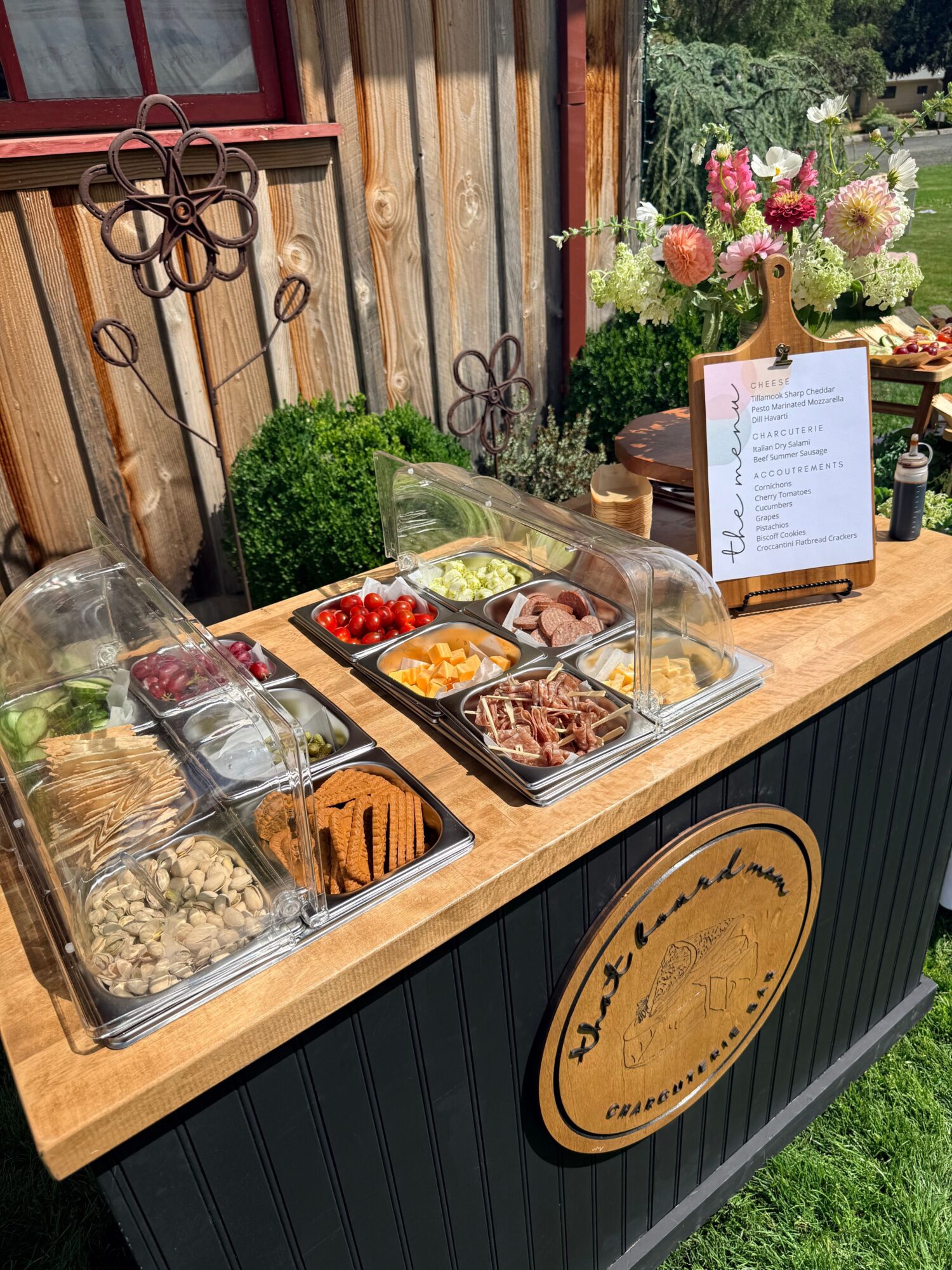 Outdoor buffet with food trays, flowers, and a menu on a wooden table, surrounded by greenery.