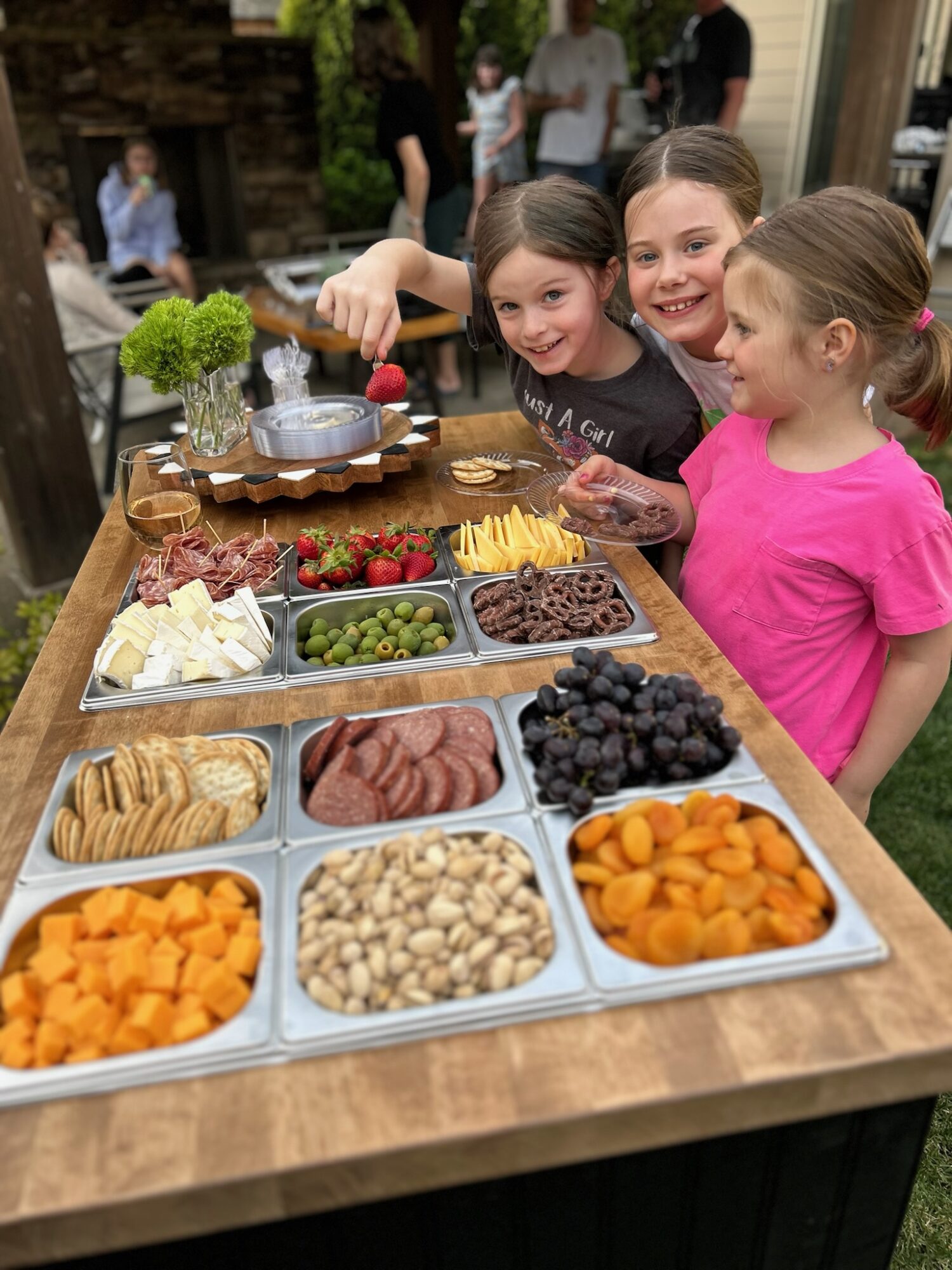 Three children smiling and pointing at a buffet of fruits, cheeses, and snacks outdoors, with other people in the background.