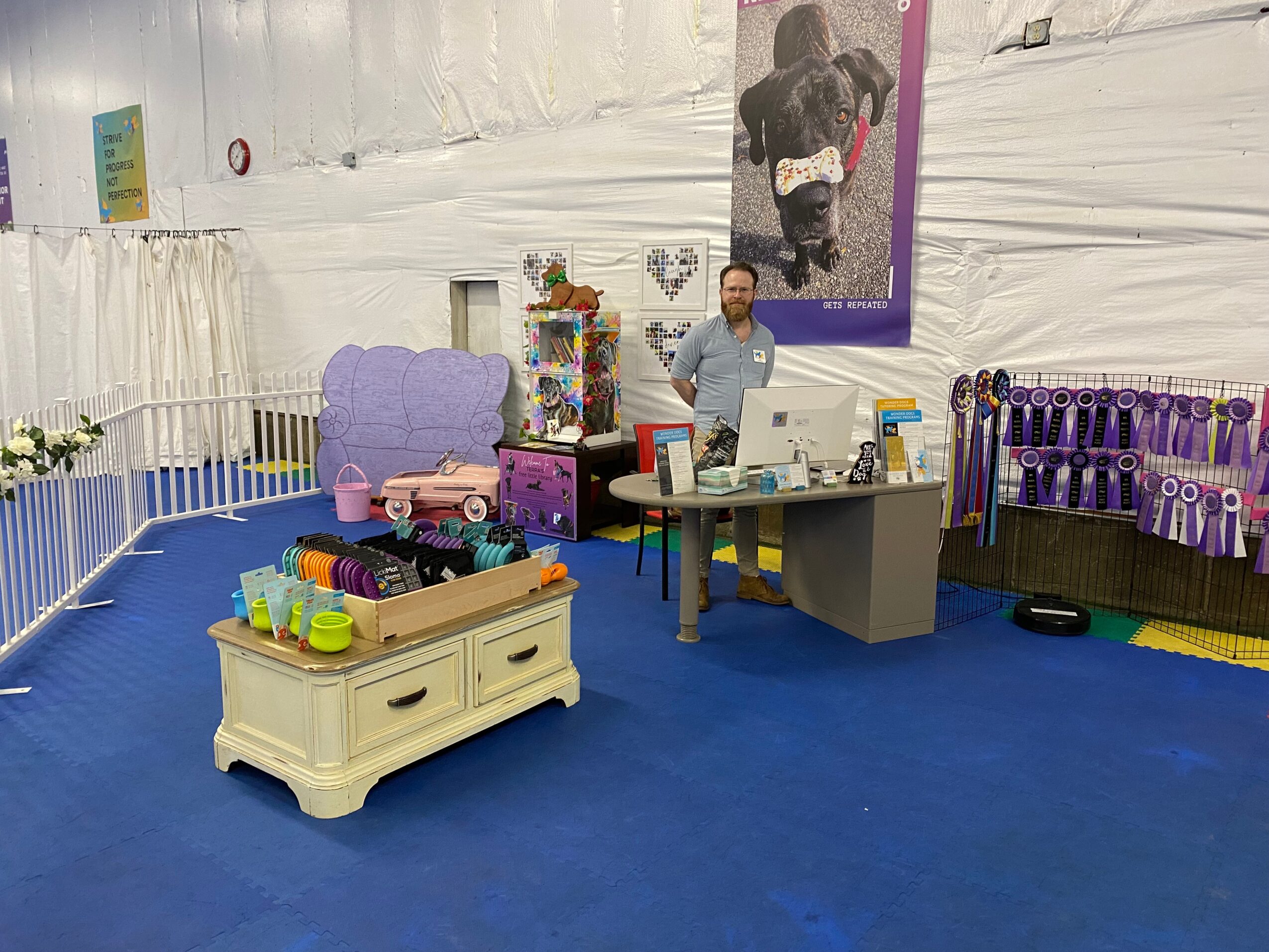 Person standing behind a table inside a tent with colorful decorations and a large mirror, blue floor, and various items on display.