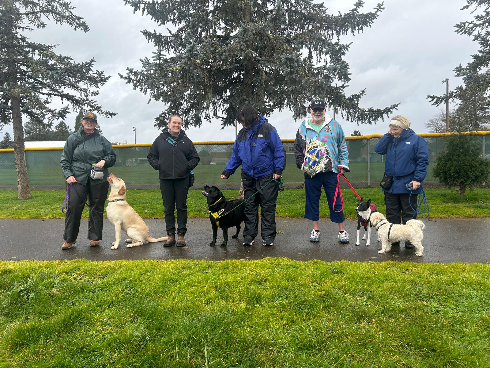 Five people with dogs standing on a paved path outdoors, trees in the background, overcast sky.