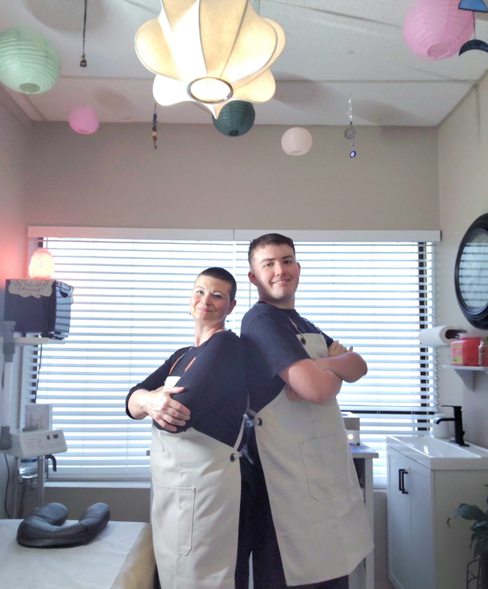 Two people stand in a room with colorful hanging lanterns and a window with blinds behind them.