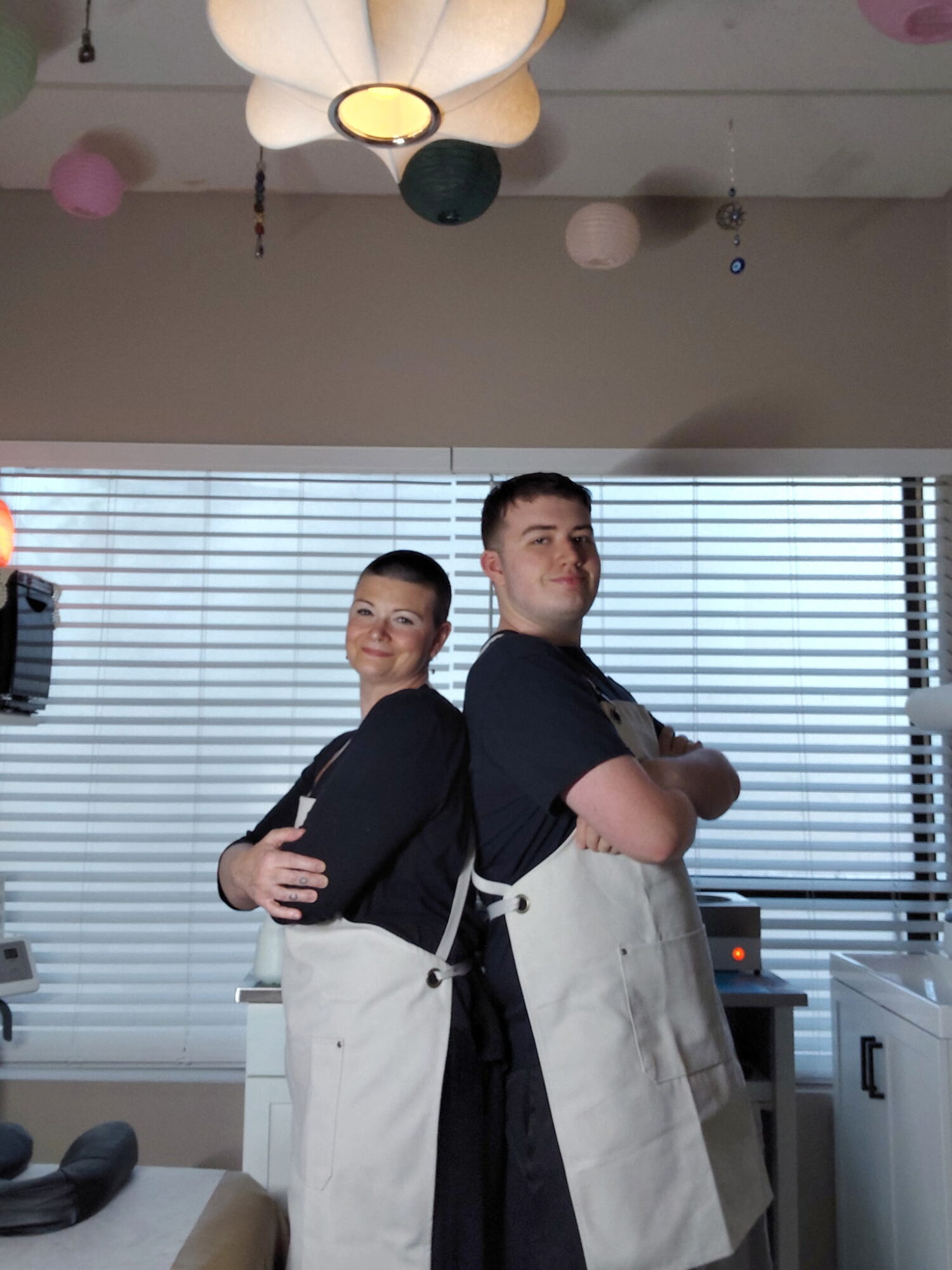 Two people stand back-to-back with arms crossed, in a room with large window blinds and hanging decorations.