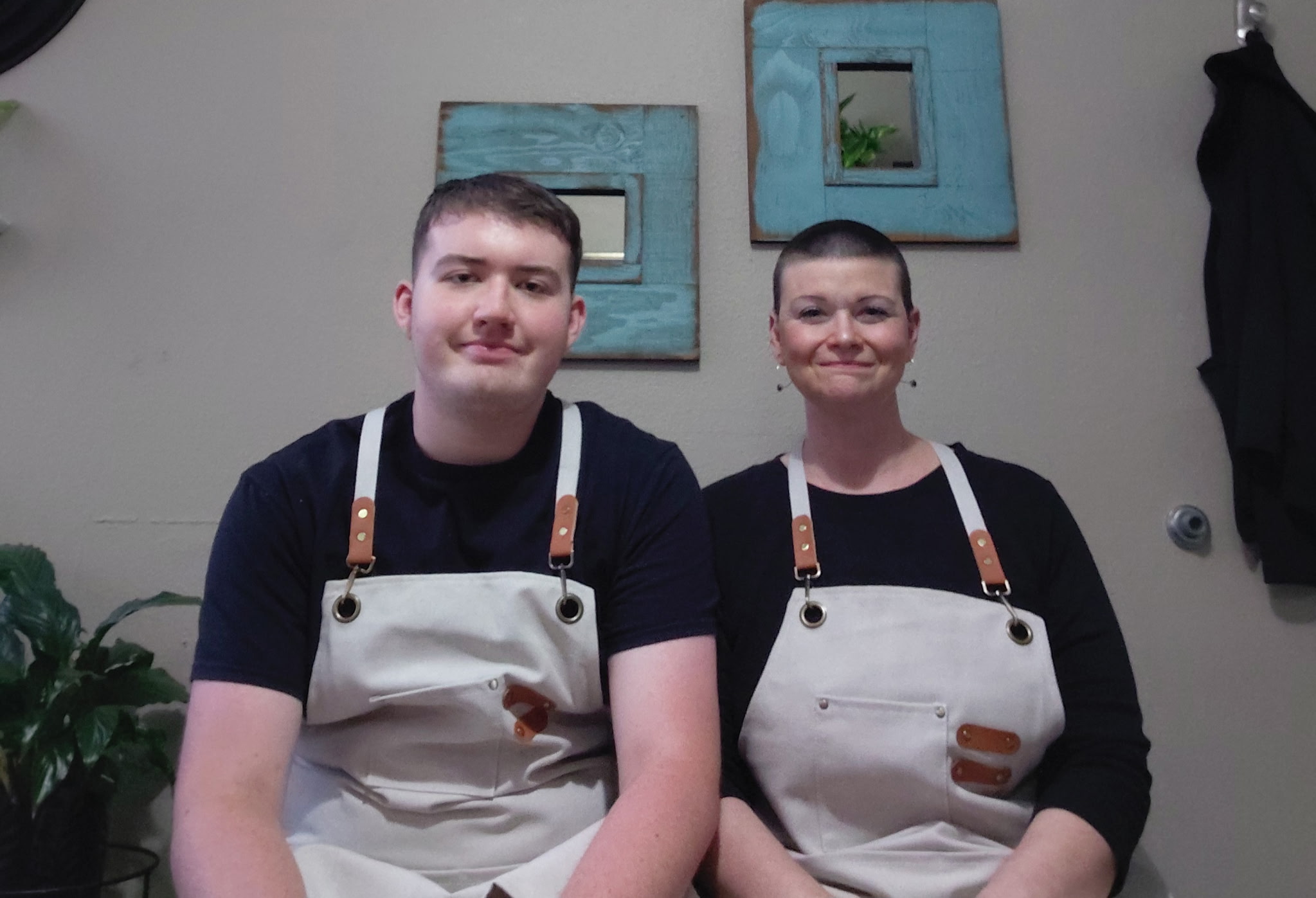 Two people wearing aprons sit side by side indoors, with framed artwork on the wall behind them.