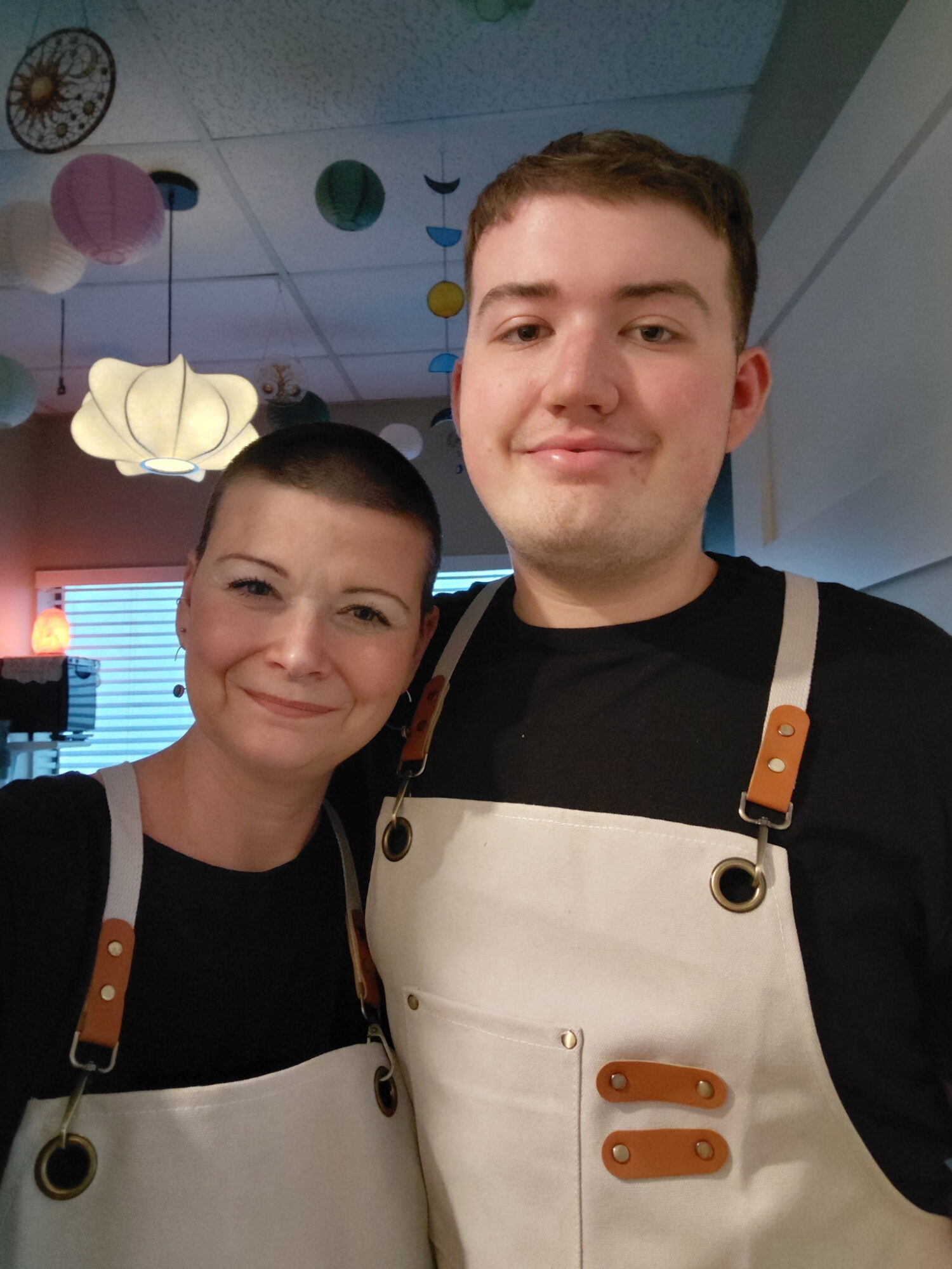 Two people wearing aprons smiling indoors, decorated with hanging paper lanterns and colorful paper balls.