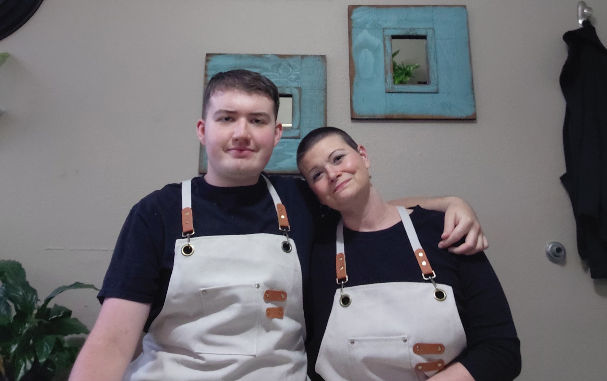Two people wearing aprons standing close together, one with arm around the other, in front of a wall with framed artwork.