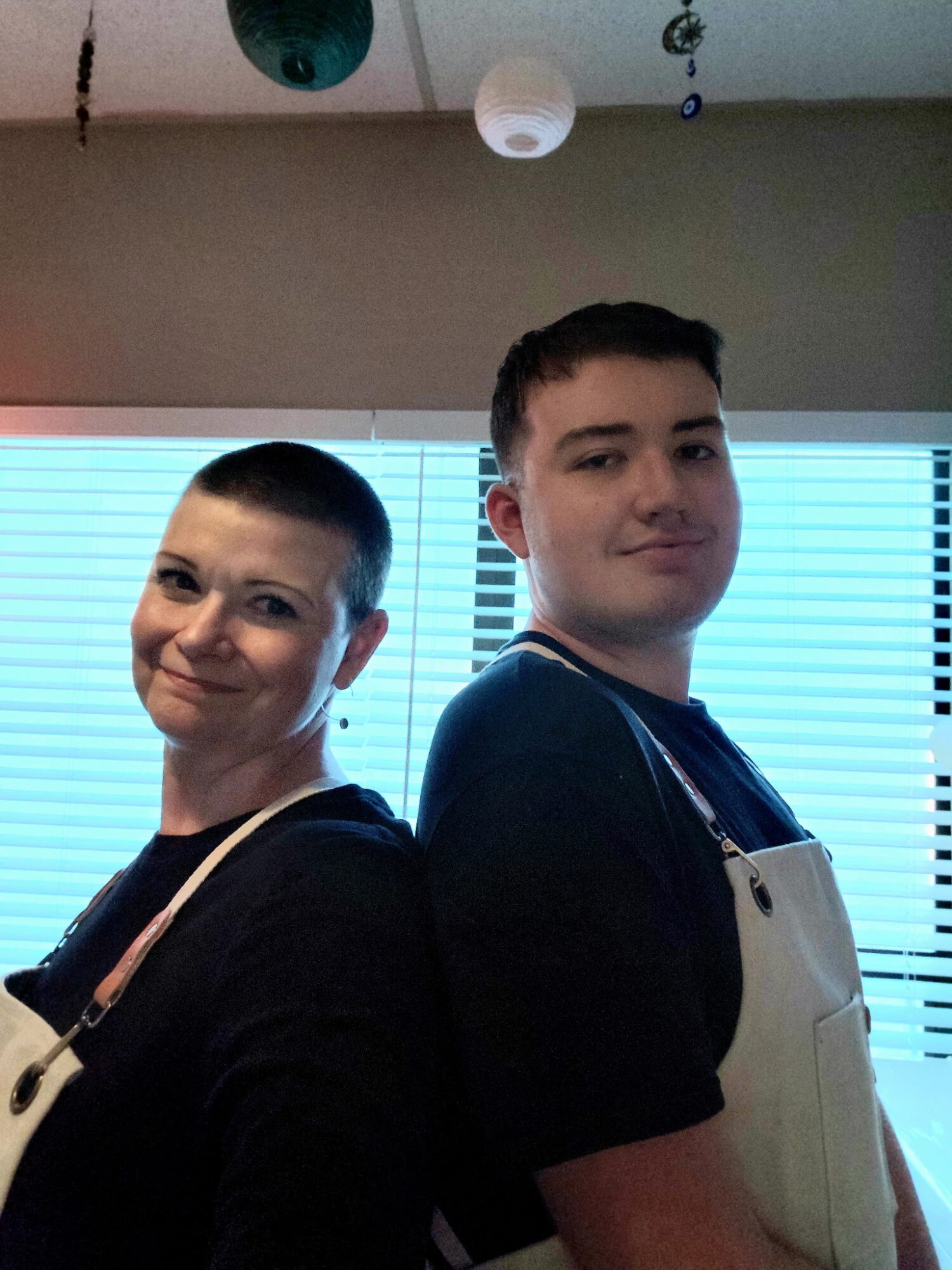Two people standing back-to-back indoors, smiling, with window blinds behind them, ceiling decorations above, one wearing an apron.
