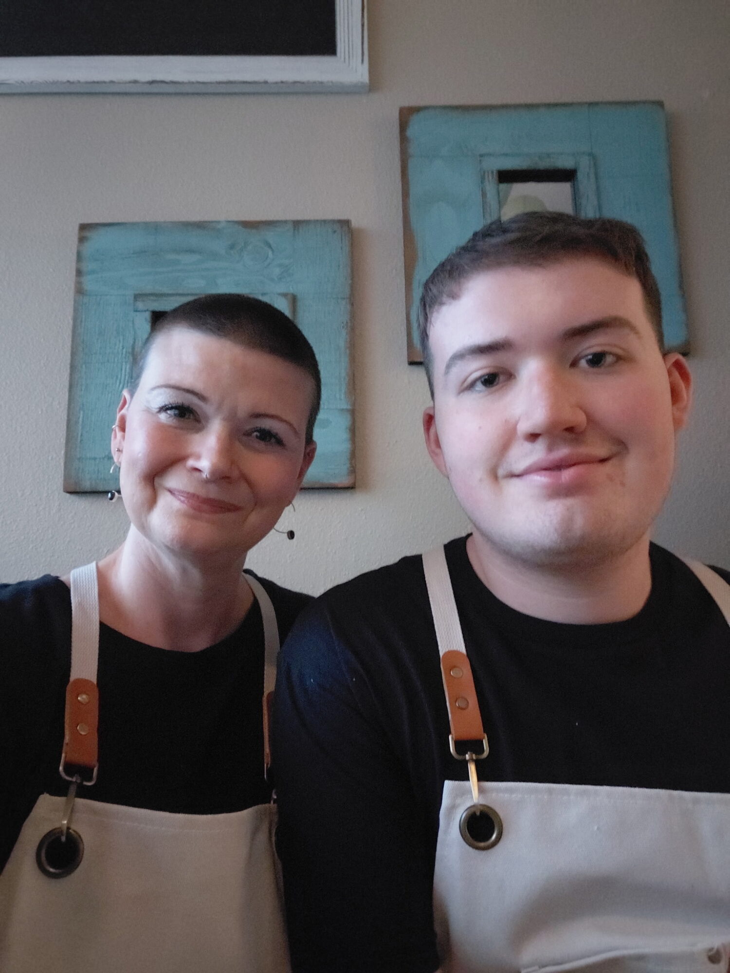 Two people smiling, wearing aprons, standing indoors with blue wall decorations behind them.