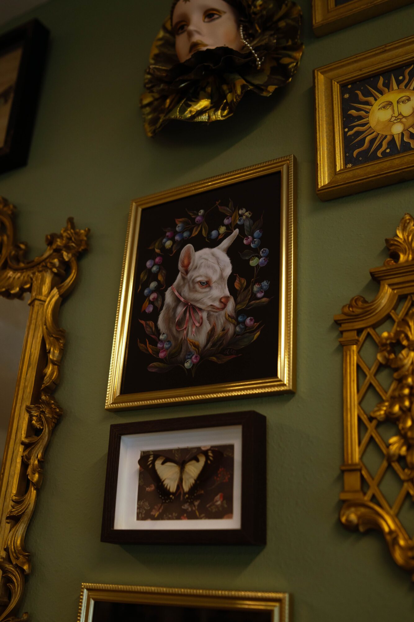 Wall decorated with framed artwork and ornate gold frames, including a pig painting and butterfly picture, with a woman's face sculpture above.