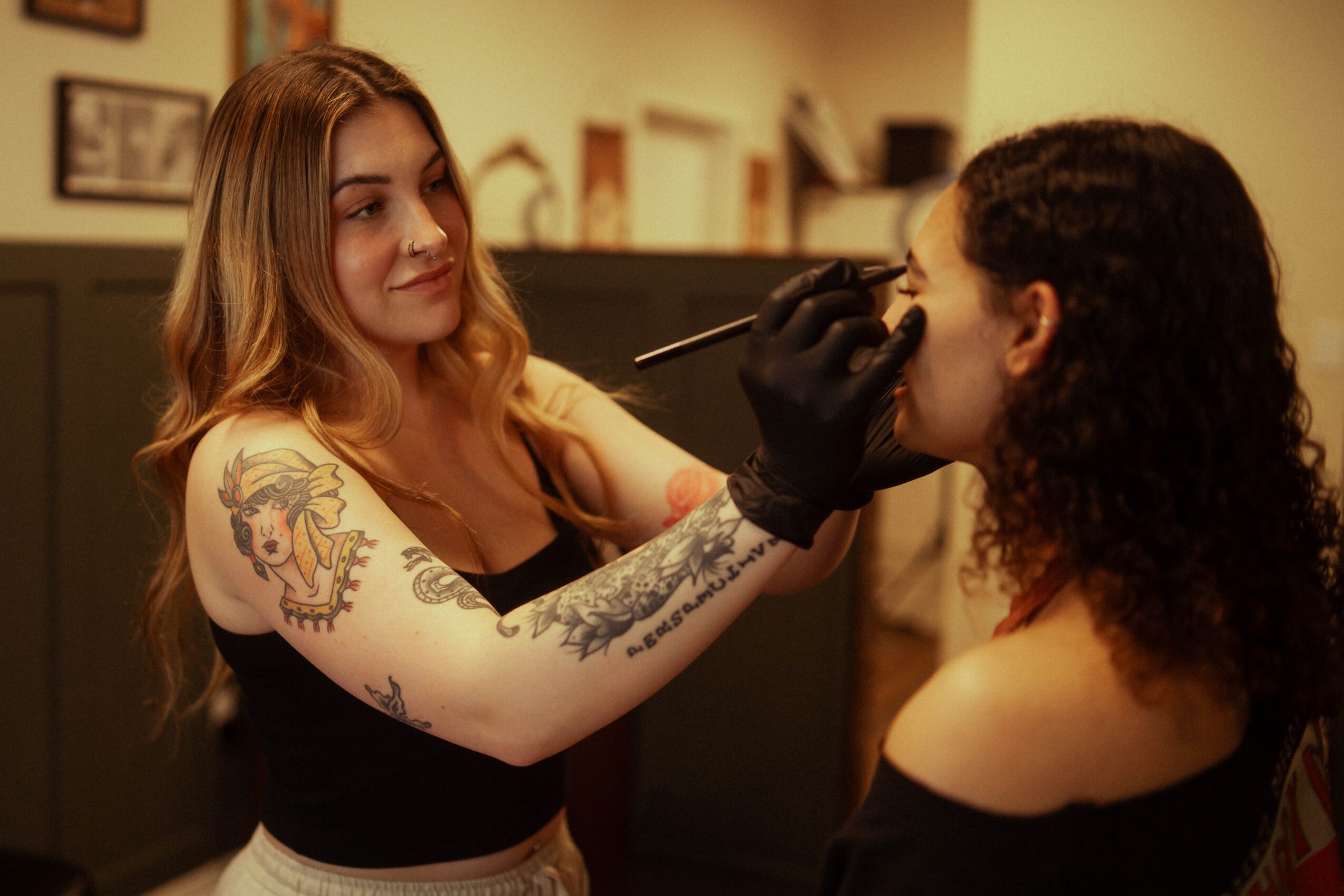 Tattooed woman applying makeup to another woman with curly hair in a room, using a brush, warm lighting.
