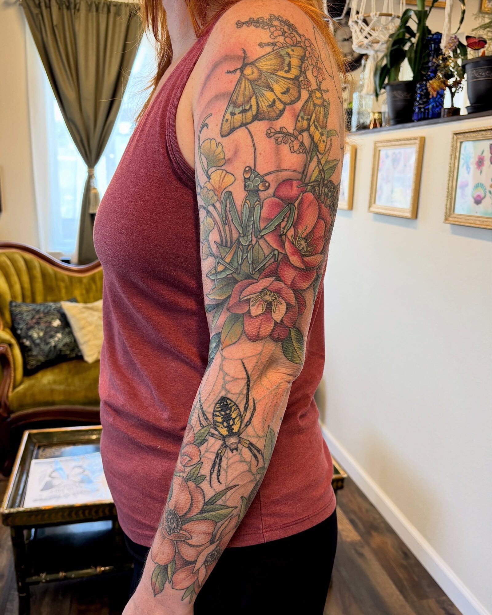 Person's arm with colorful floral and insect tattoo, wearing a sleeveless top, in a room with framed pictures and a sofa.