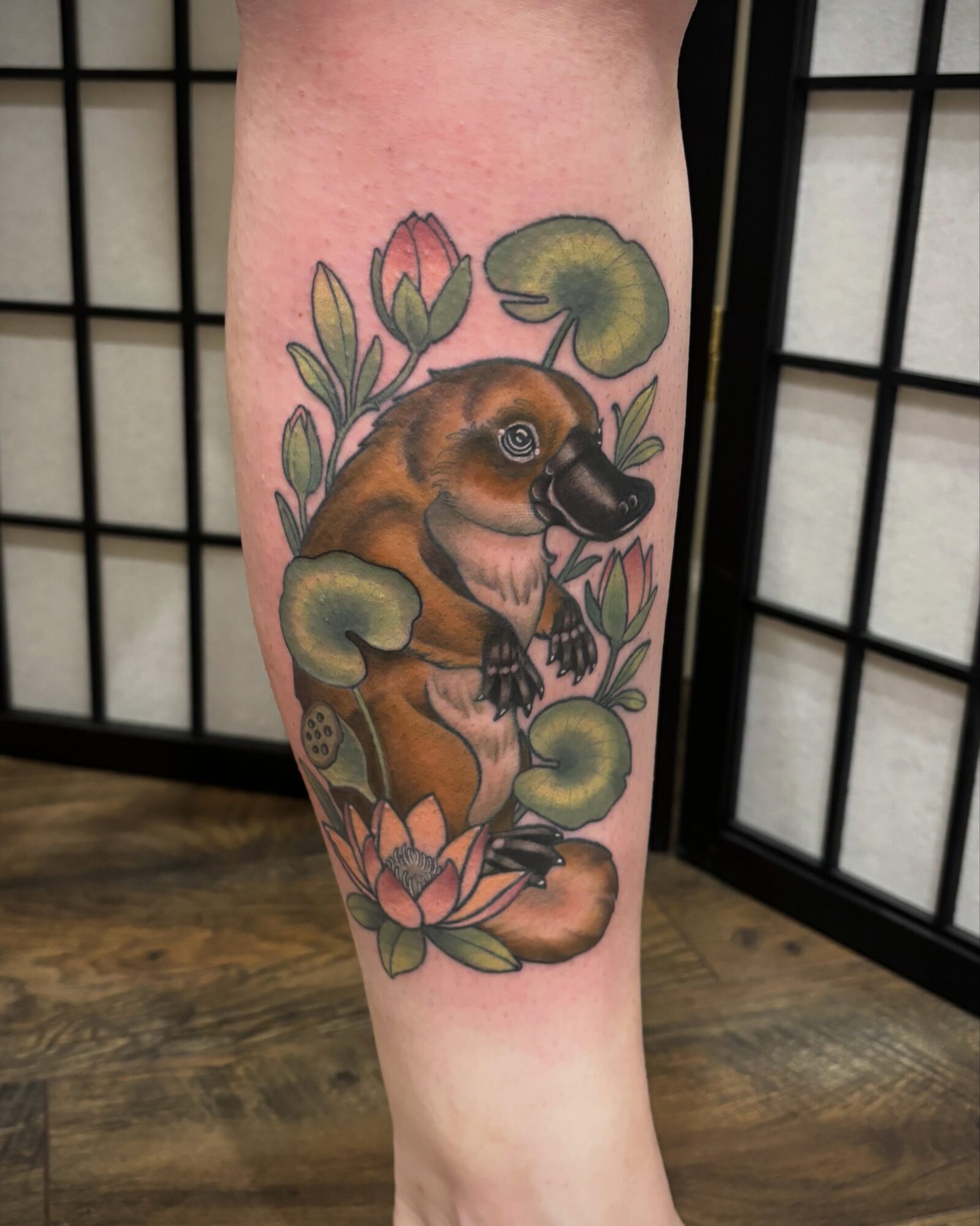 Tattoo of a baby otter surrounded by lotus flowers and lily pads on a person's leg.