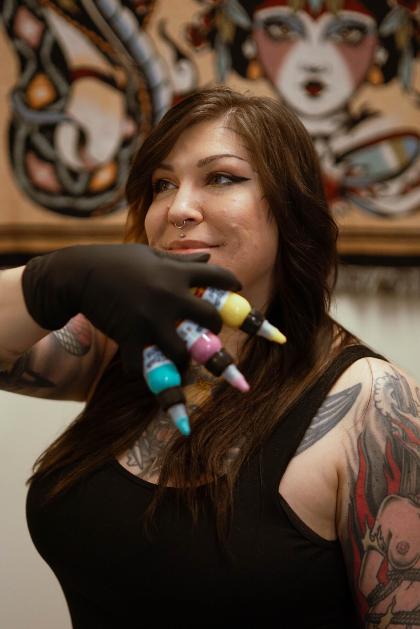 Tattooed woman with long hair getting a colorful tattoo on her shoulder, background features a mural with animal faces.