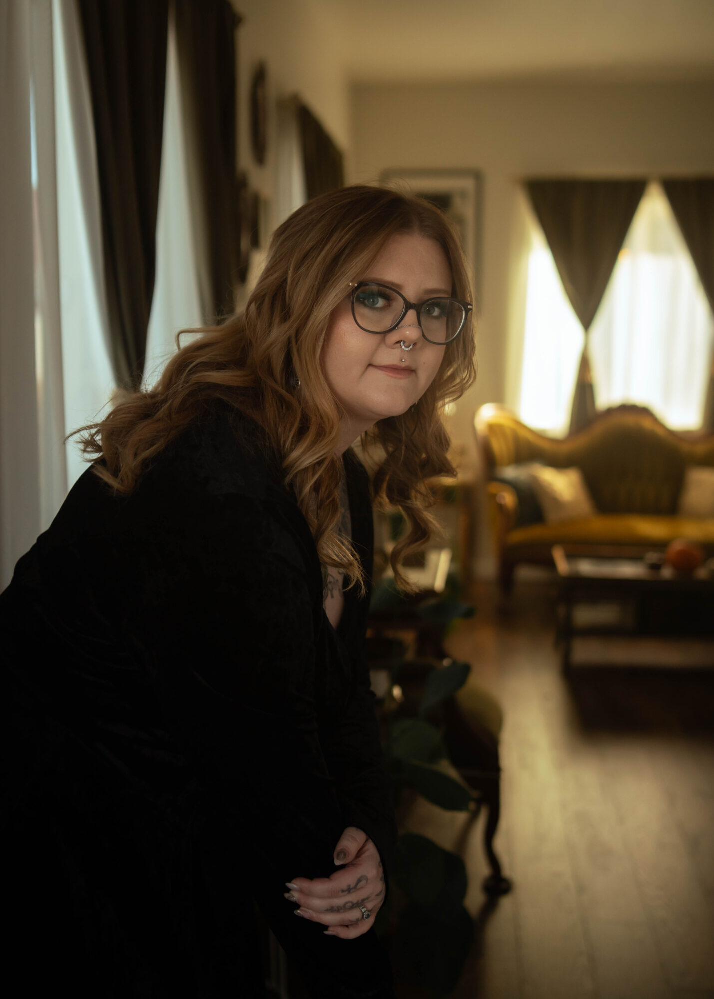 Woman with glasses and wavy hair sitting in a living room with curtains and furniture behind her.