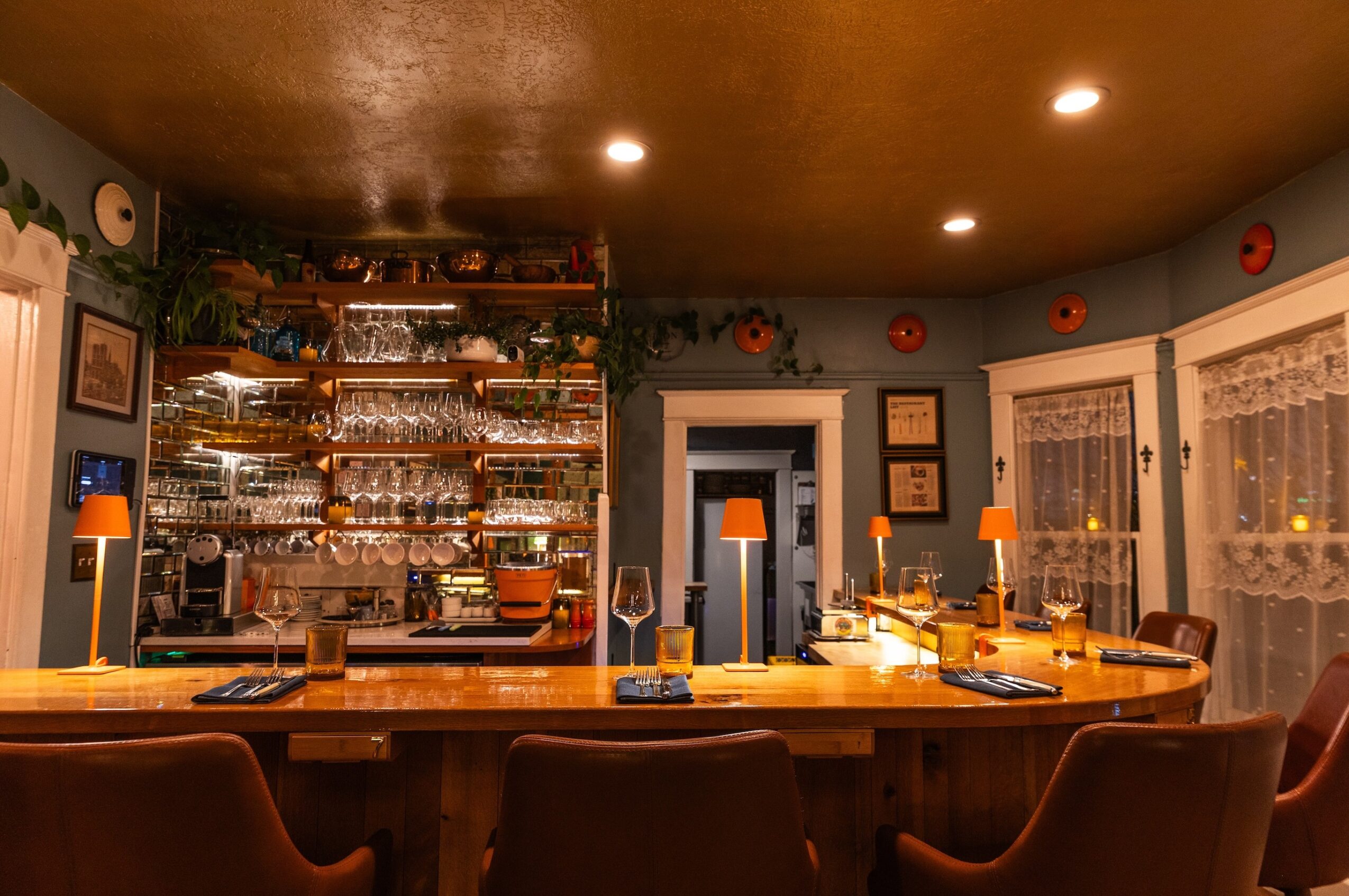Cozy dining area with a large wooden table, bar shelves filled with glasses, and warm lighting, windows with lace curtains.