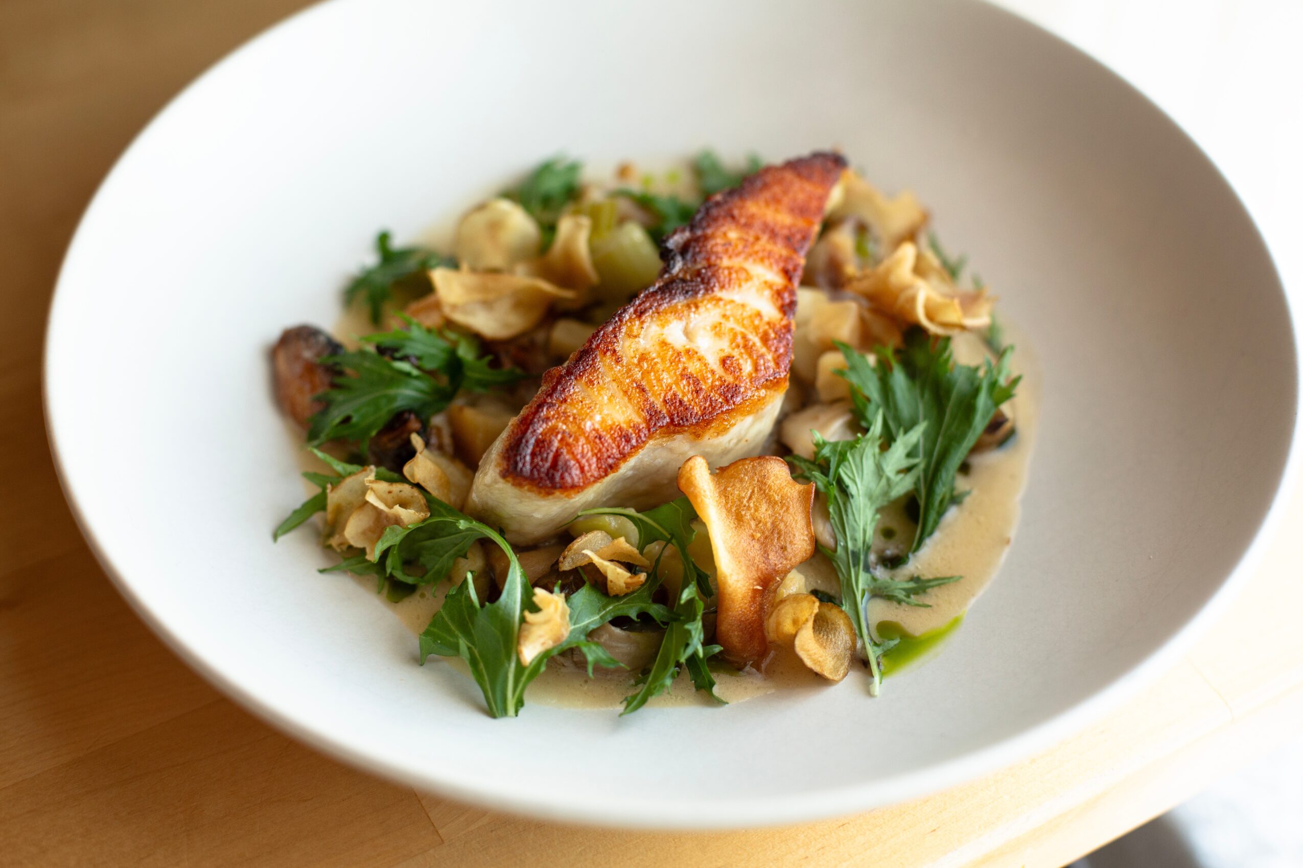 A white bowl contains a cooked fish fillet, pasta, and green herbs, served in a light sauce.