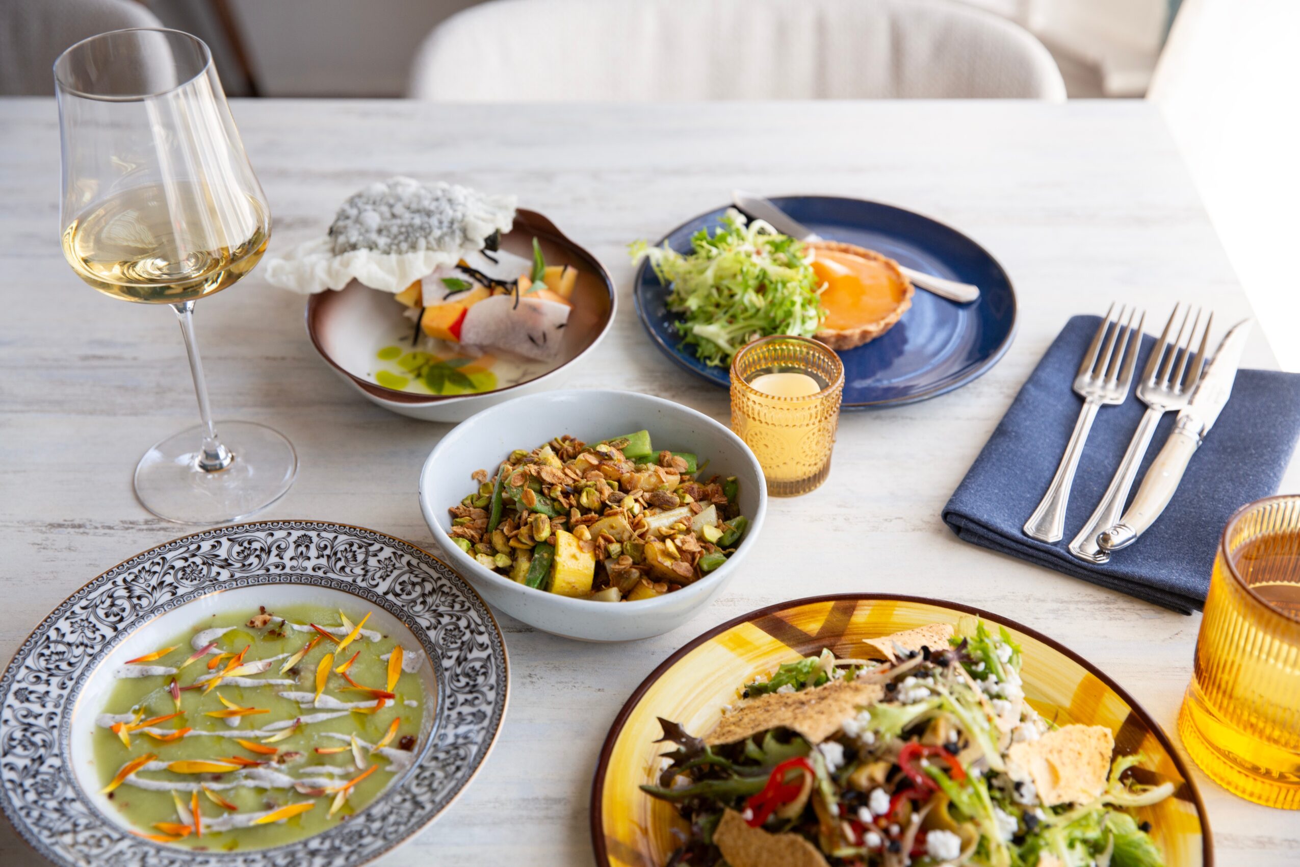 Table with various dishes, a glass of white wine, and utensils on a white tablecloth.
