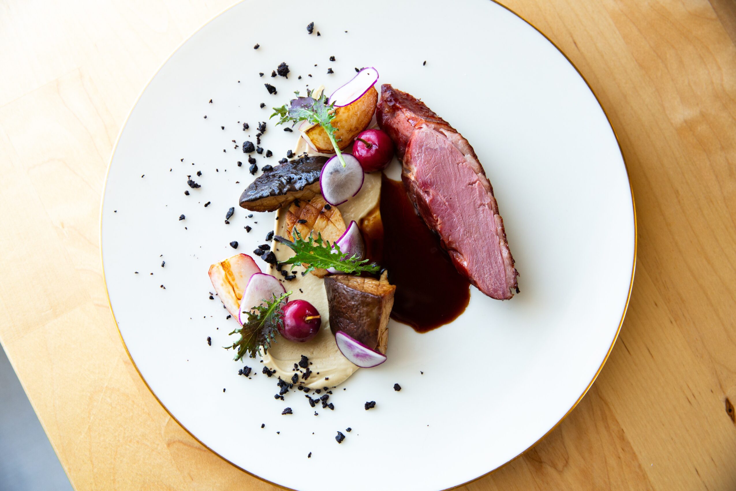 Plate with sliced cooked meat, vegetables, and sauce, garnished with herbs and black crumbs, on a wooden surface.
